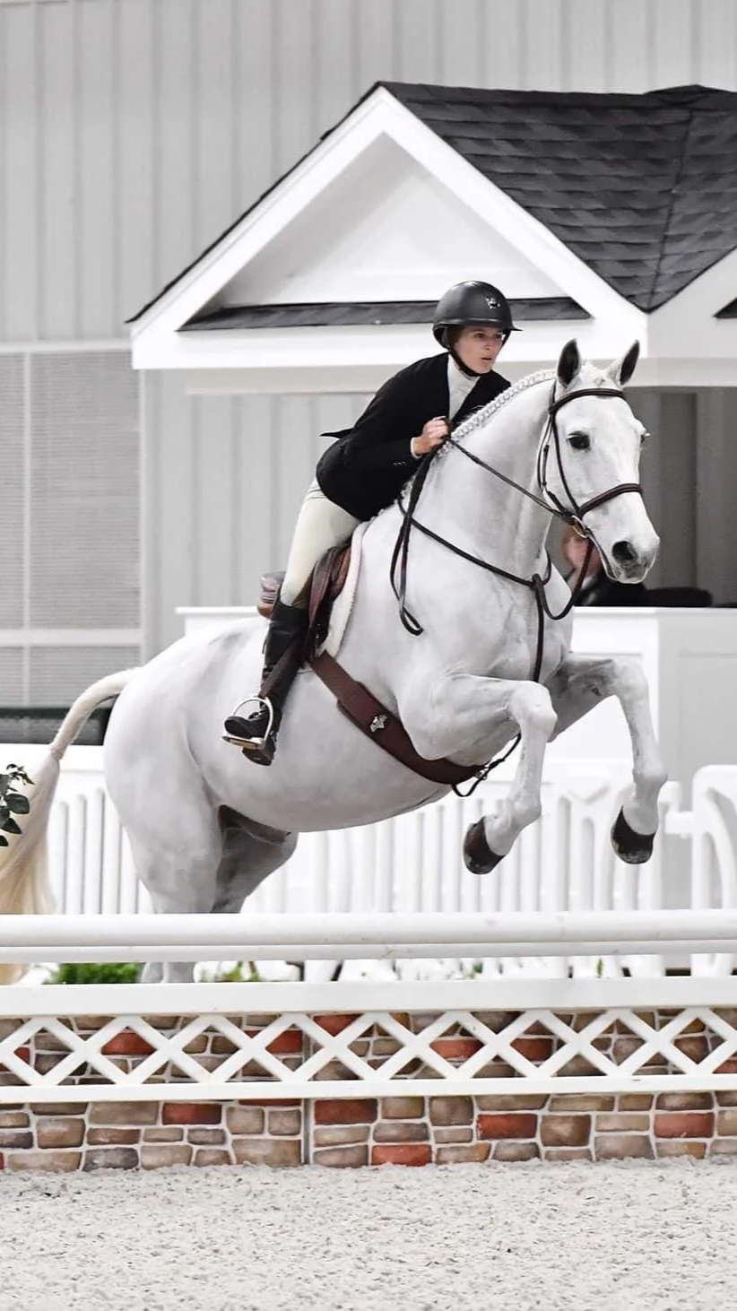 A rider dressed in black and white equestrian attire riding a white horse during an indoor show jumping competition, with a decorative backdrop resembling a house.