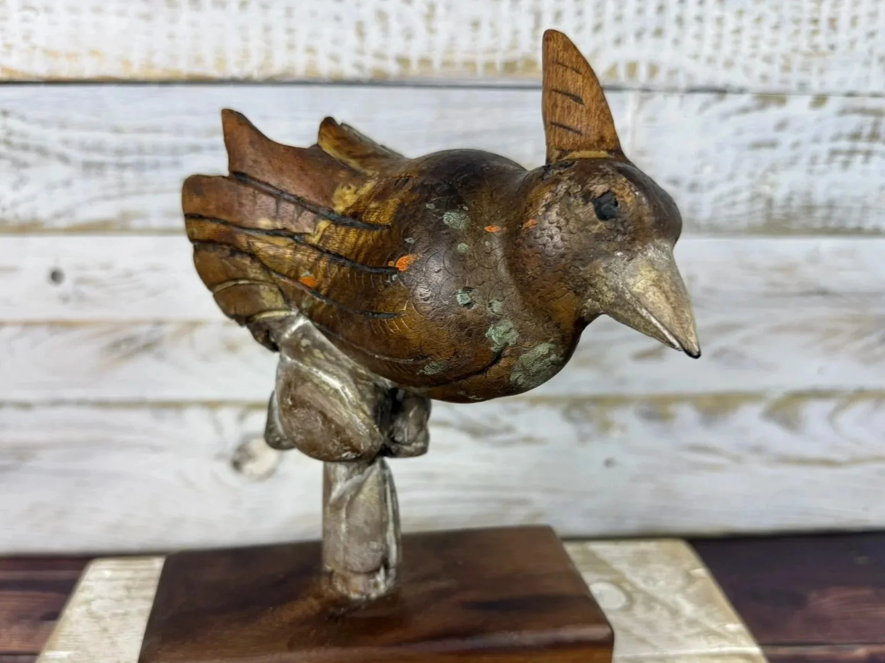 Wooden sculpture of a bird perched on a small branch, with a light-colored wooden background.