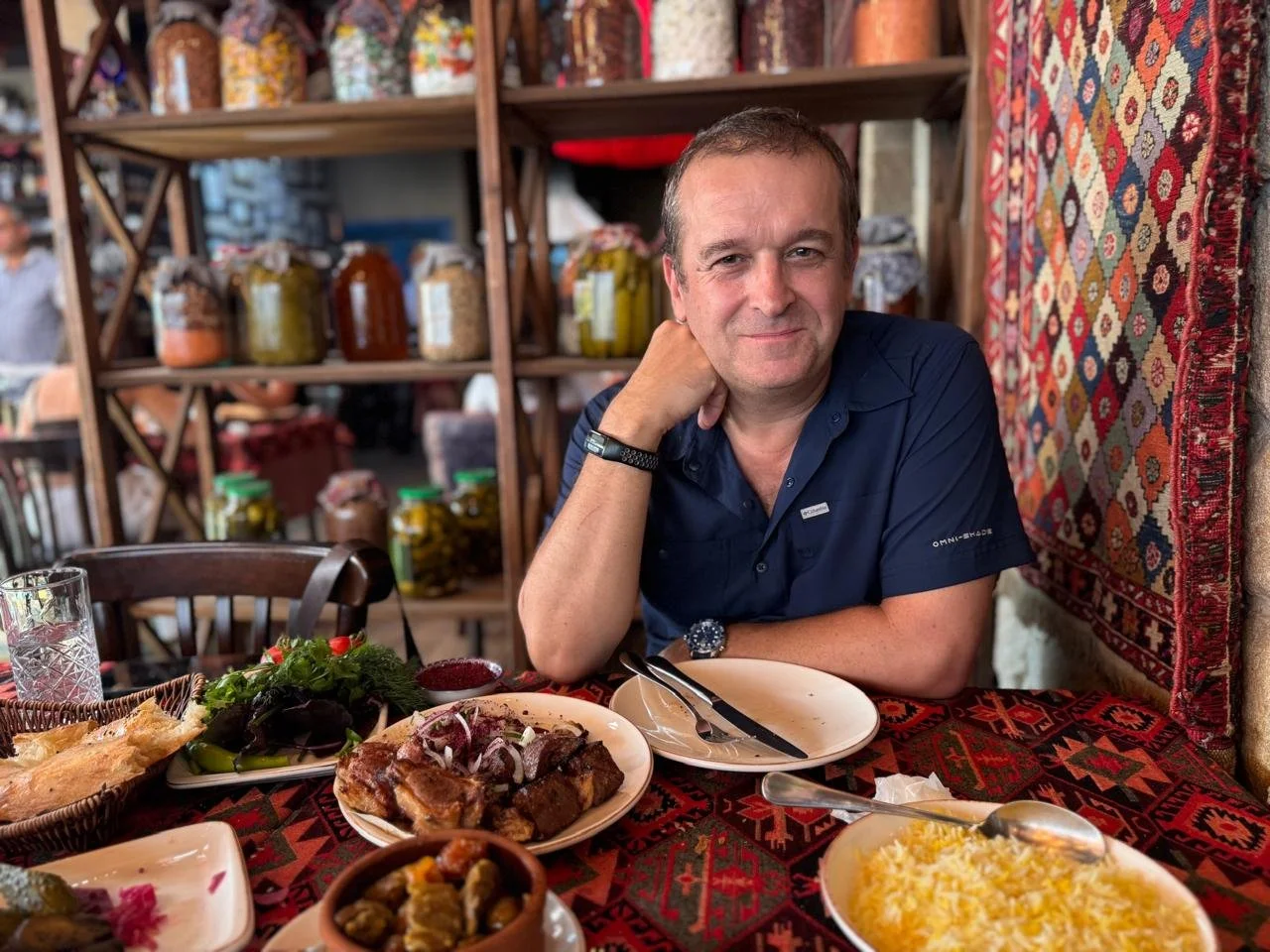 A man sitting at a table in a restaurant with traditional patterned textiles on the wall, with plates of food including a salad, grilled meat, rice, and appetizers, and jars of preserved vegetables in the background.