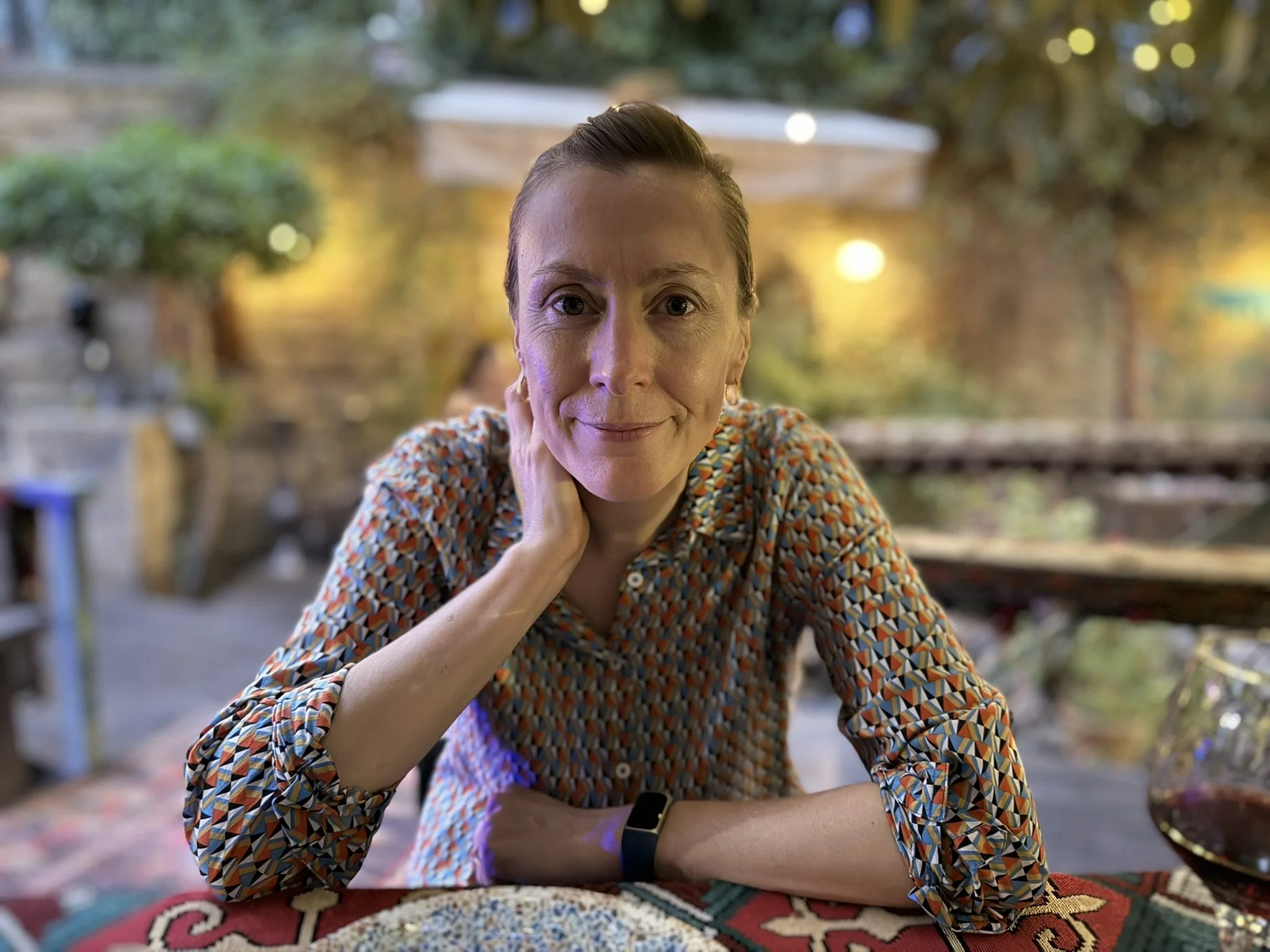 A woman with short hair and fair skin sitting at a table, resting her head on her left hand, looking at the camera and smiling. She is wearing a colorful geometric patterned shirt. The background is blurred with outdoor lighting and greenery.