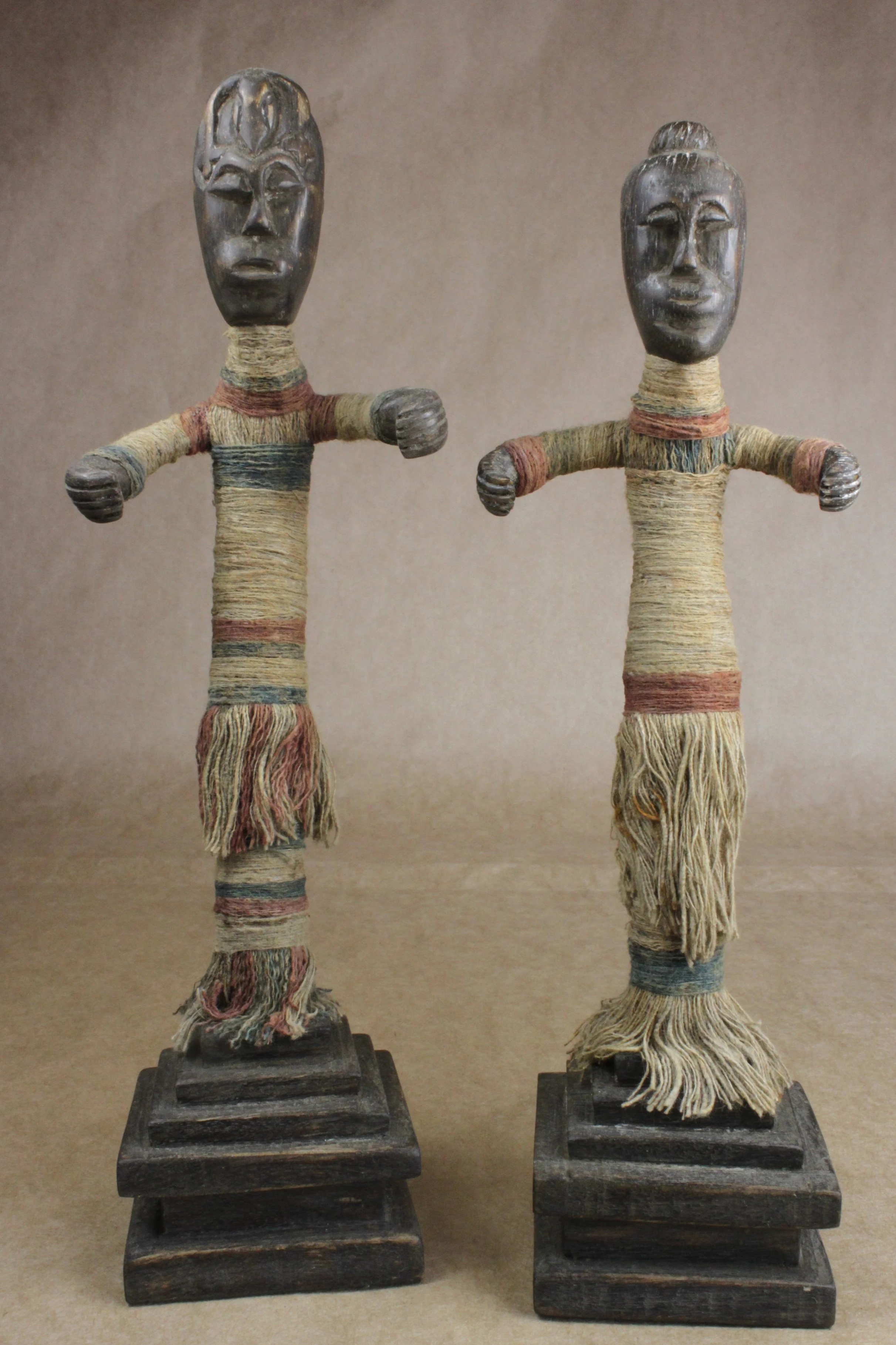 Two tribal figurines with cloth-wrapped bodies and dark carved faces, standing on black rectangular bases.