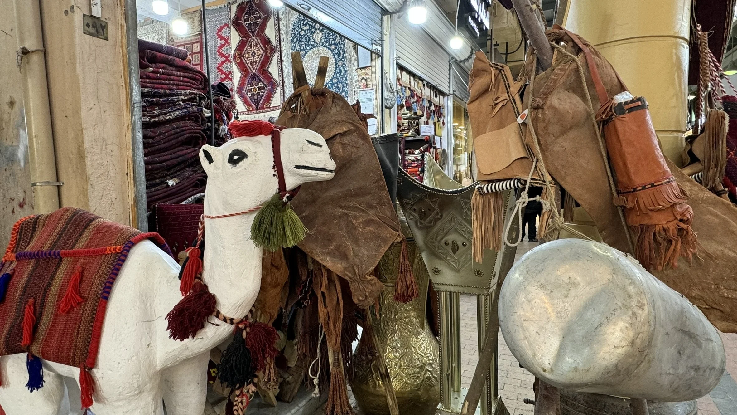 A storefront displaying vintage leather bags, woven rugs, and a decorative camel sculpture with saddle and tassels in an indoor market.