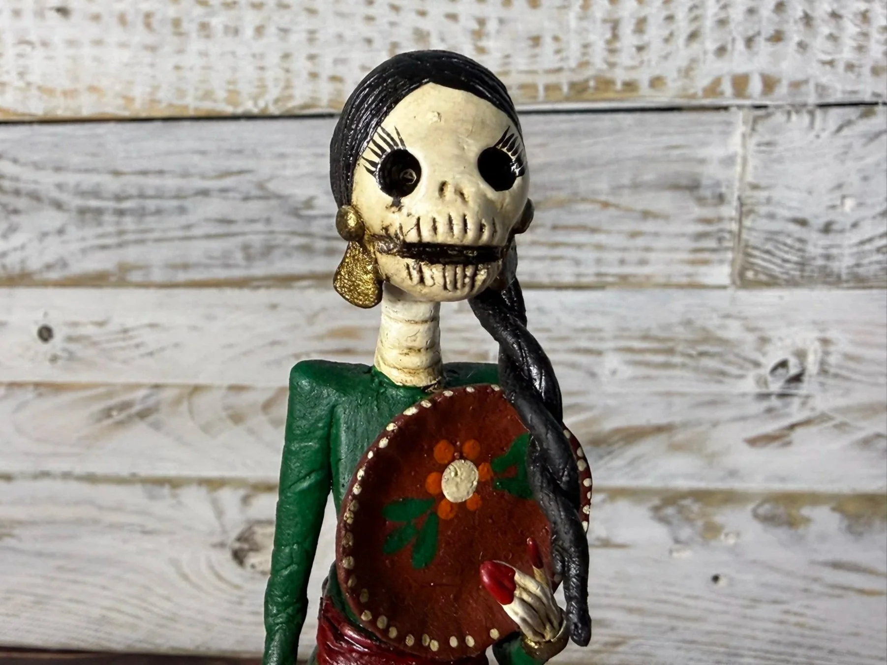 Decorative sculpture of a woman with a skull face, black hair, and a green dress, holding a red shield with floral designs, set against a weathered wooden background.