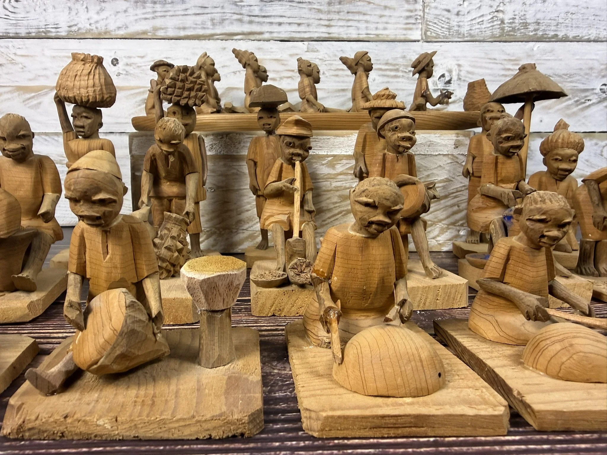 Wooden carved Yoruba thorn wood figurines depicting a group of people with some holding umbrellas, seated and standing, with a background of wooden plank wall.
