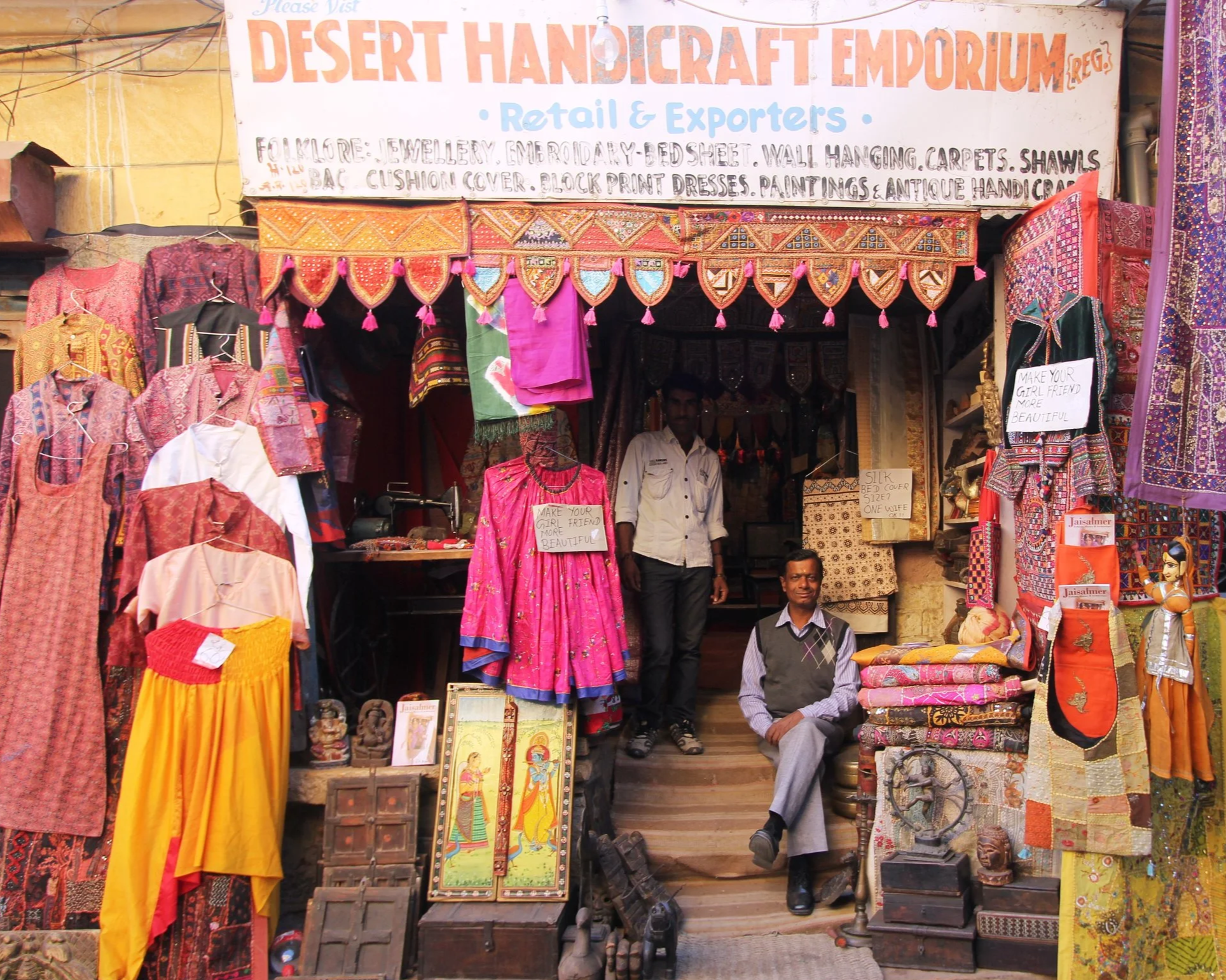 A colorful market stall with a sign that reads "Desert Handicraft Emporium". The stall displays various traditional textiles, garments, and decorative items, with two men sitting and standing inside. Items include embroidered clothes, shawls, paintings, and antique artifacts.