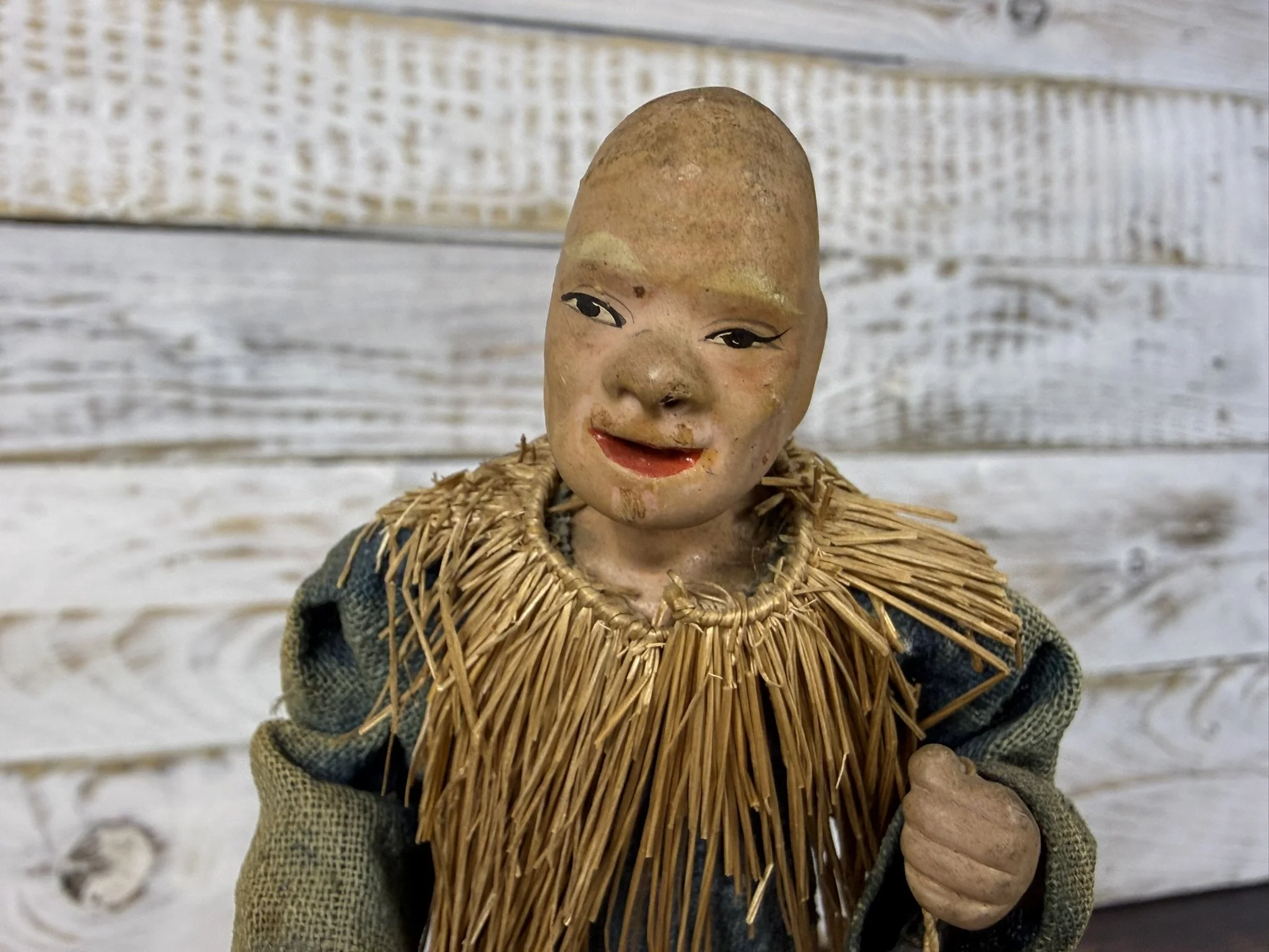 Close-up of an old wooden doll with a painted face, wearing a straw collar and a green fabric garment, set against a background of weathered white wooden planks.