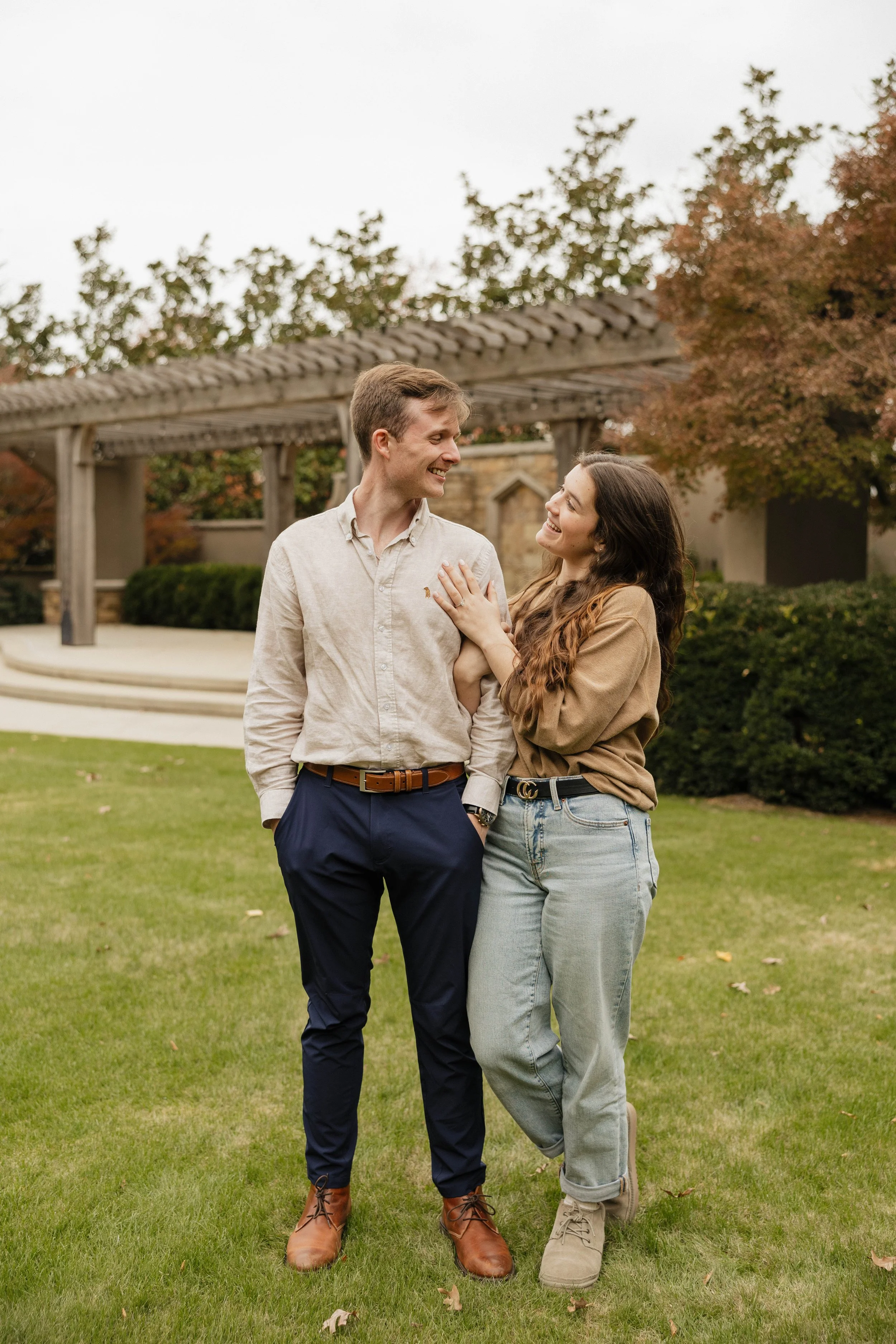 A young man and woman standing on a grassy area, smiling at each other, outdoors in front of a wooden structure and trees with autumn leaves.