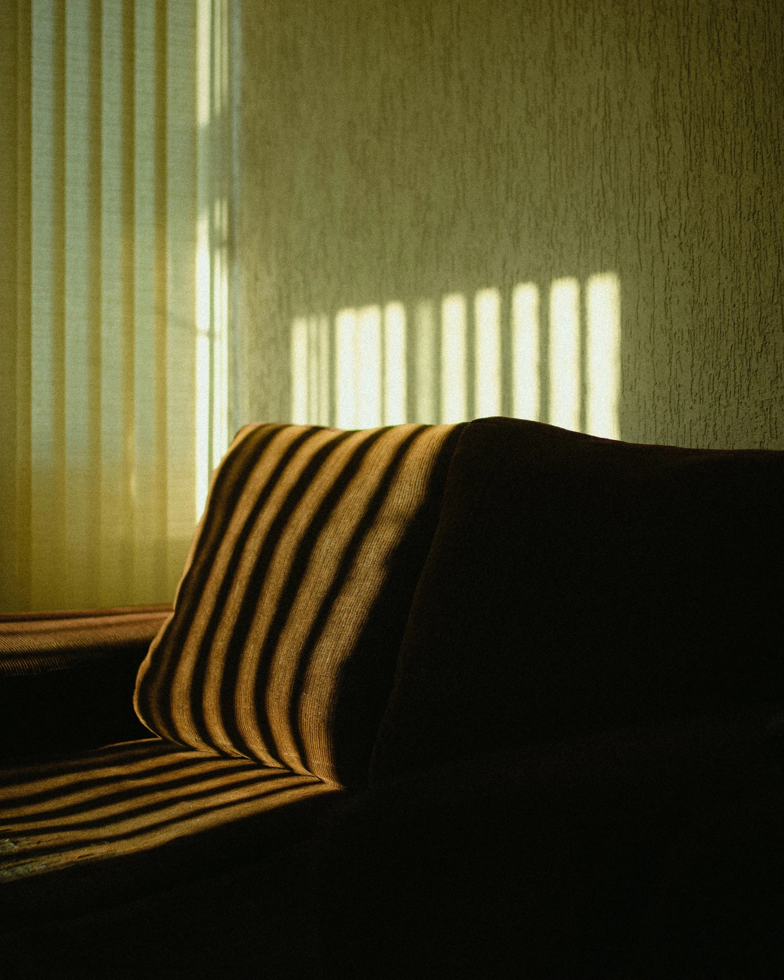 A striped cushion on a dark brown sofa with sunlight casting shadows through window blinds onto the wall and sofa.