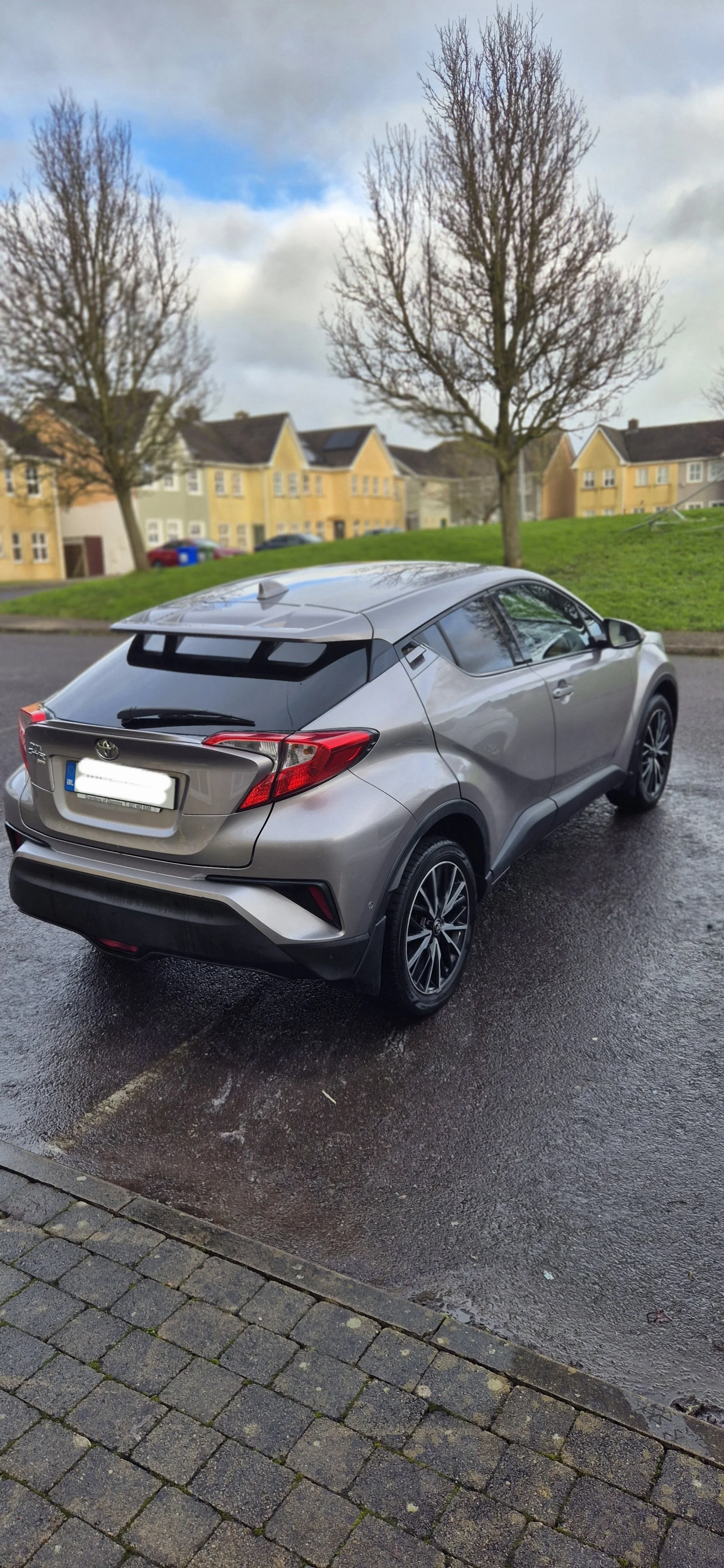 A silver Toyota C-HR parked on a wet street with a grassy area and leafless trees behind houses under a cloudy sky.