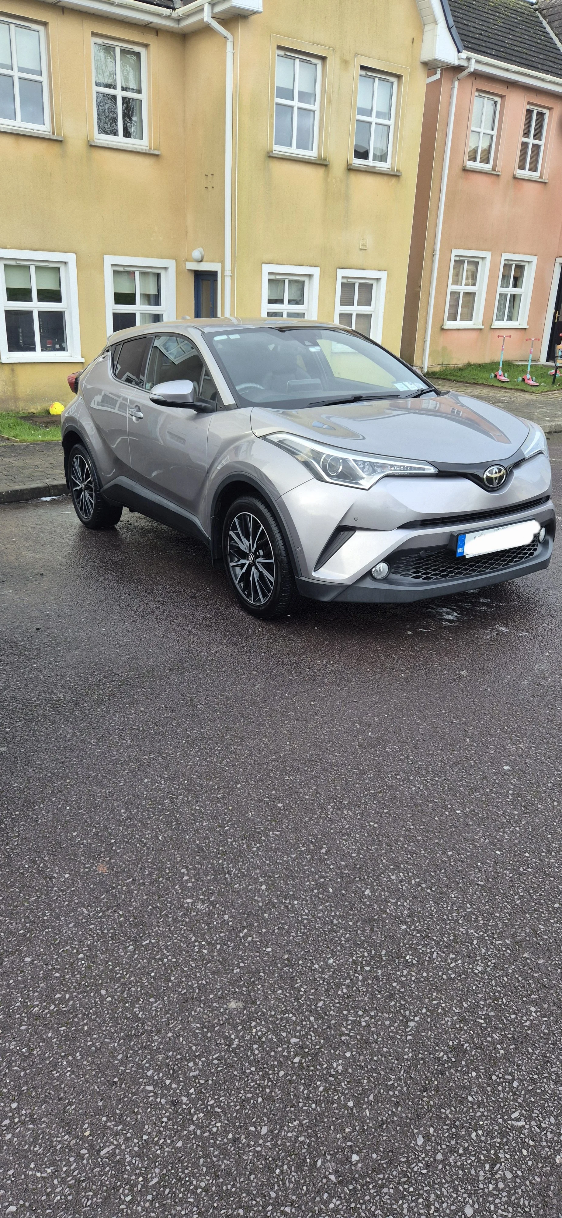 A silver Toyota C-HR parked on a wet asphalt street in front of colorful residential buildings with yellow and pink facades.