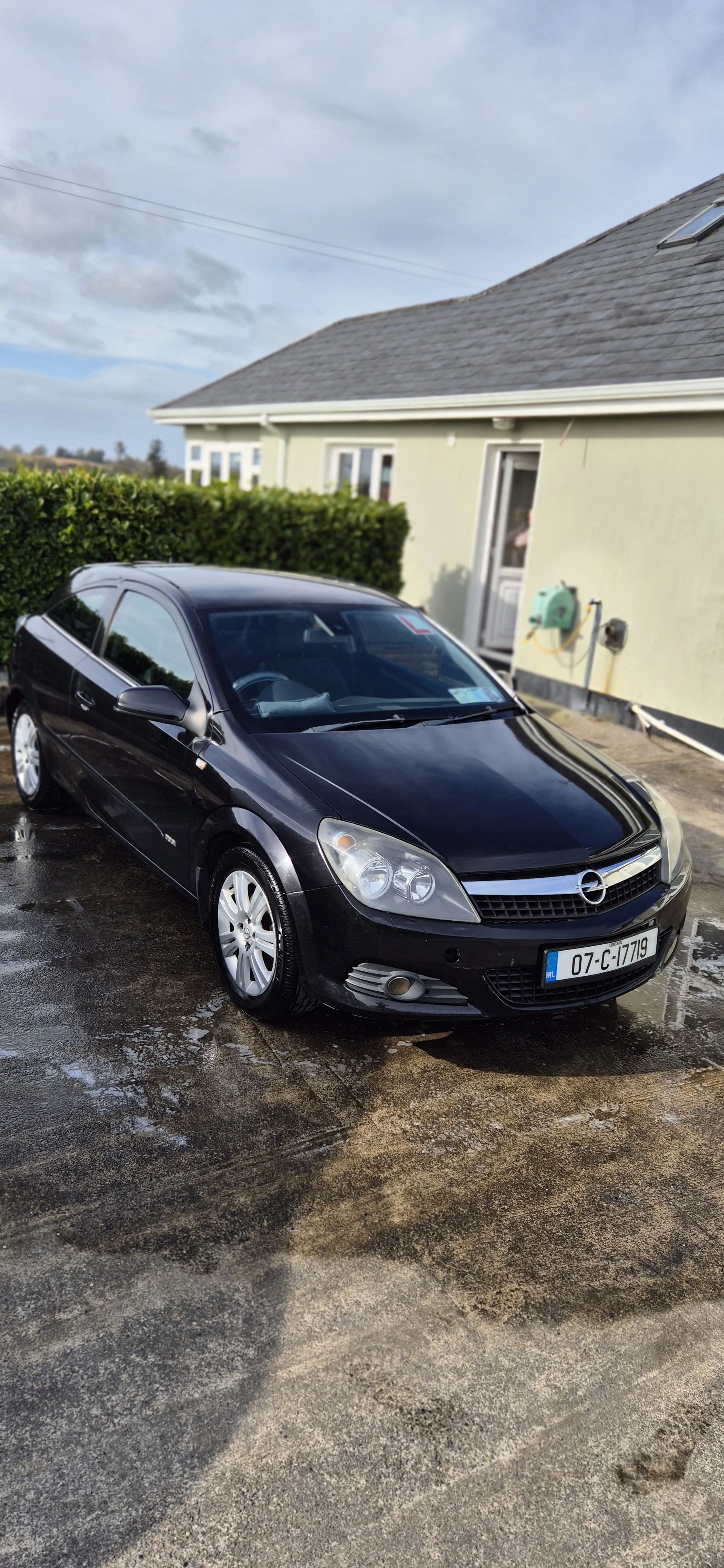 A black Opel Astra parked outside a house on a wet driveway, with grey clouds overhead.