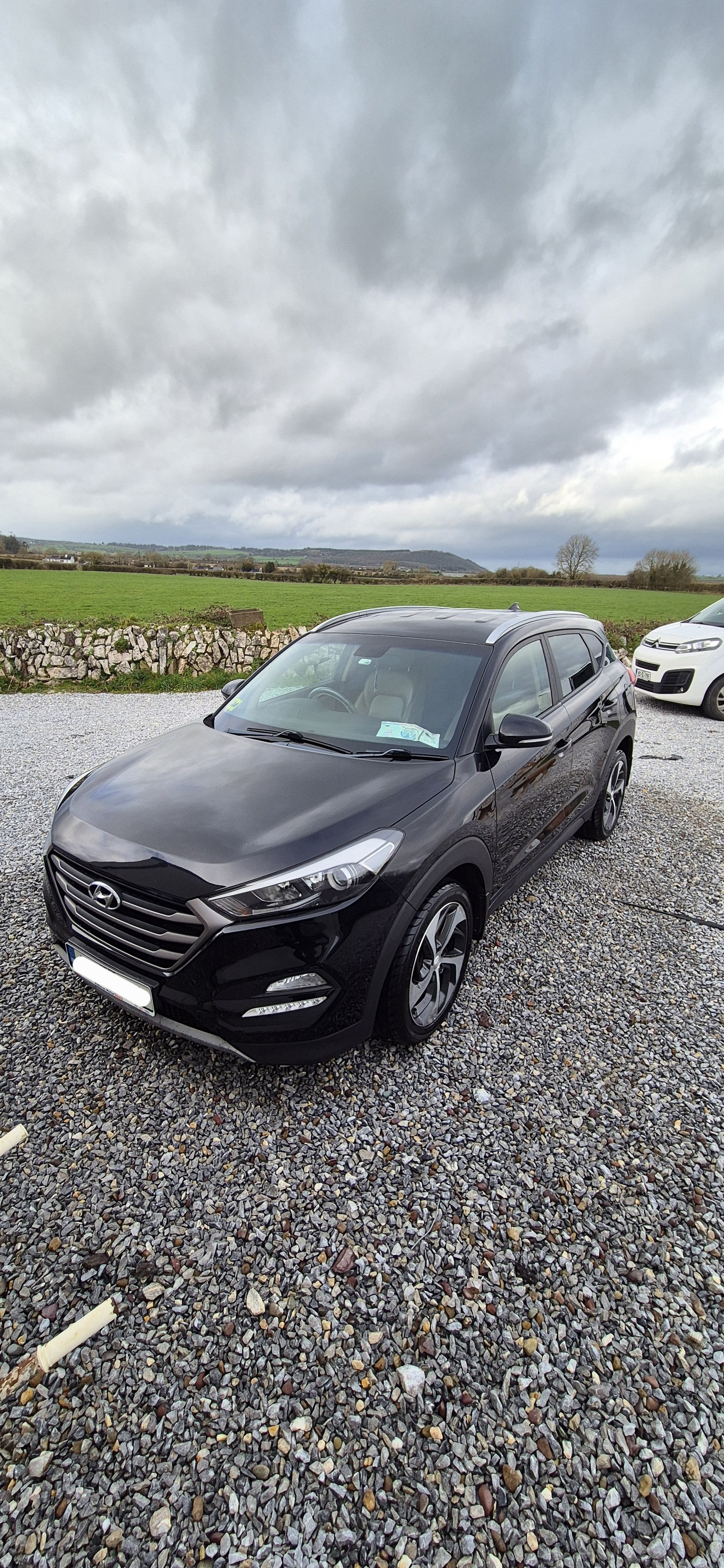 A black Hyundai SUV parked on a gravel lot with green fields and cloudy sky in the background.
