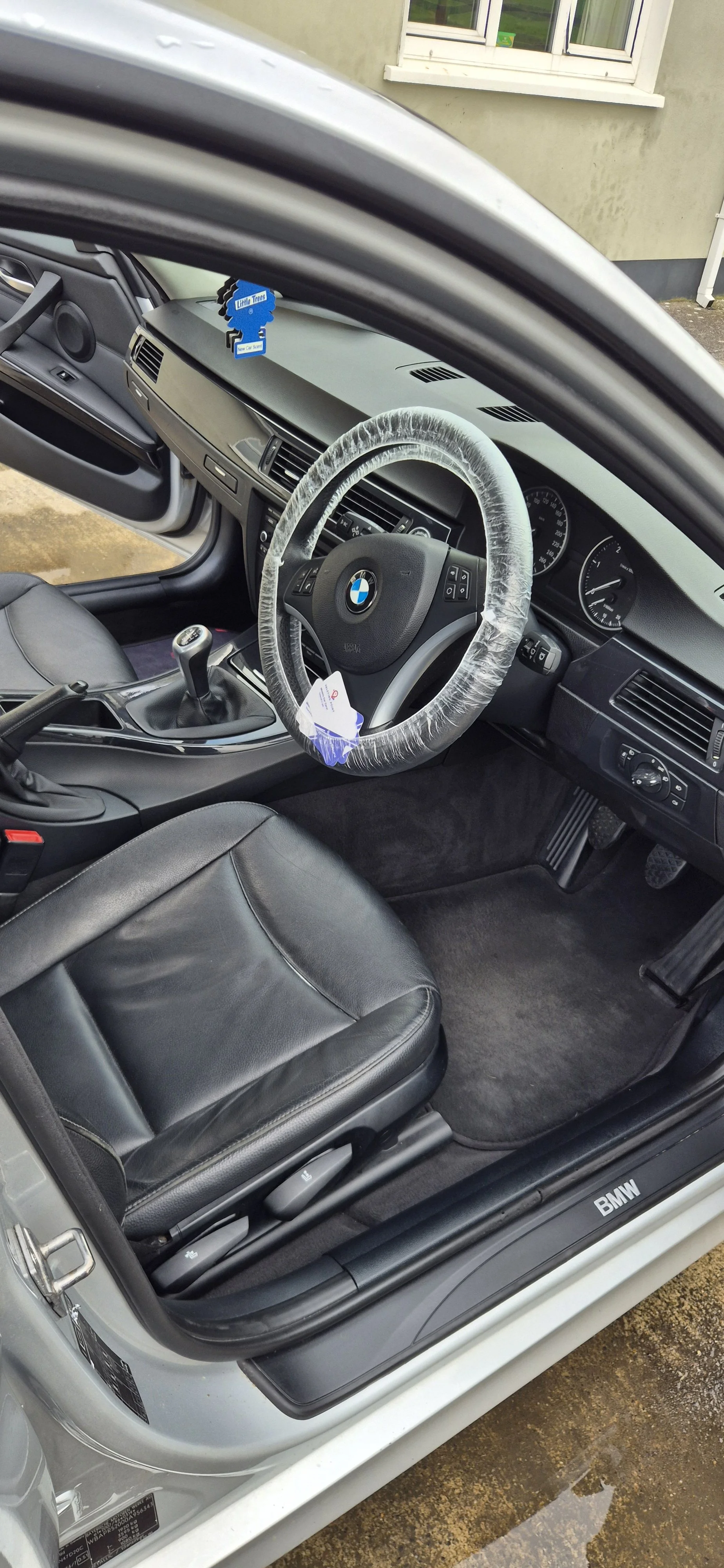Interior of a BMW car showing the driver's seat, steering wheel with a protective cover, gear shift, dashboard, and front window with a house outside.