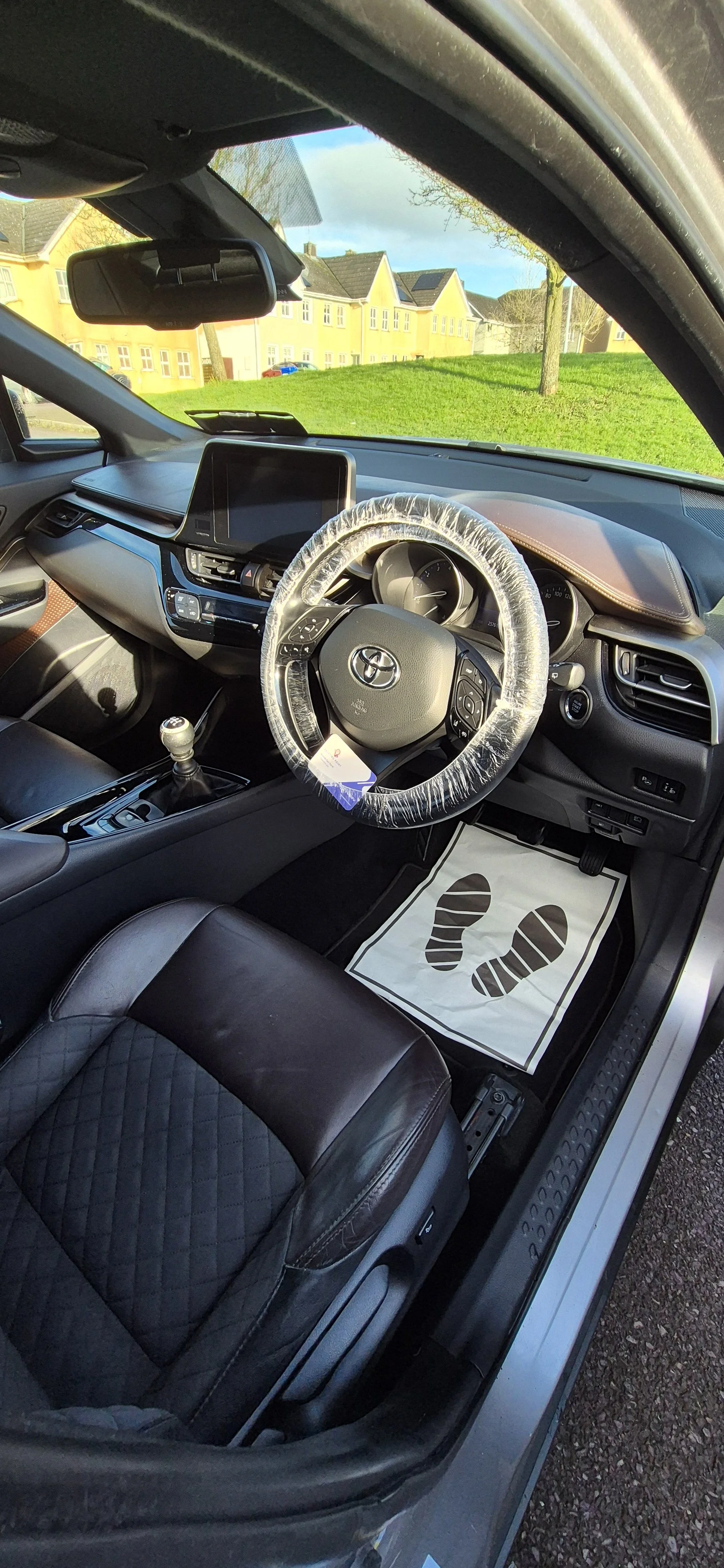 Interior of a Toyota car showing the steering wheel wrapped with a clear plastic cover, a gear shift, a black leather seat with a quilted pattern, and a floor mat with black footprints on a white background.