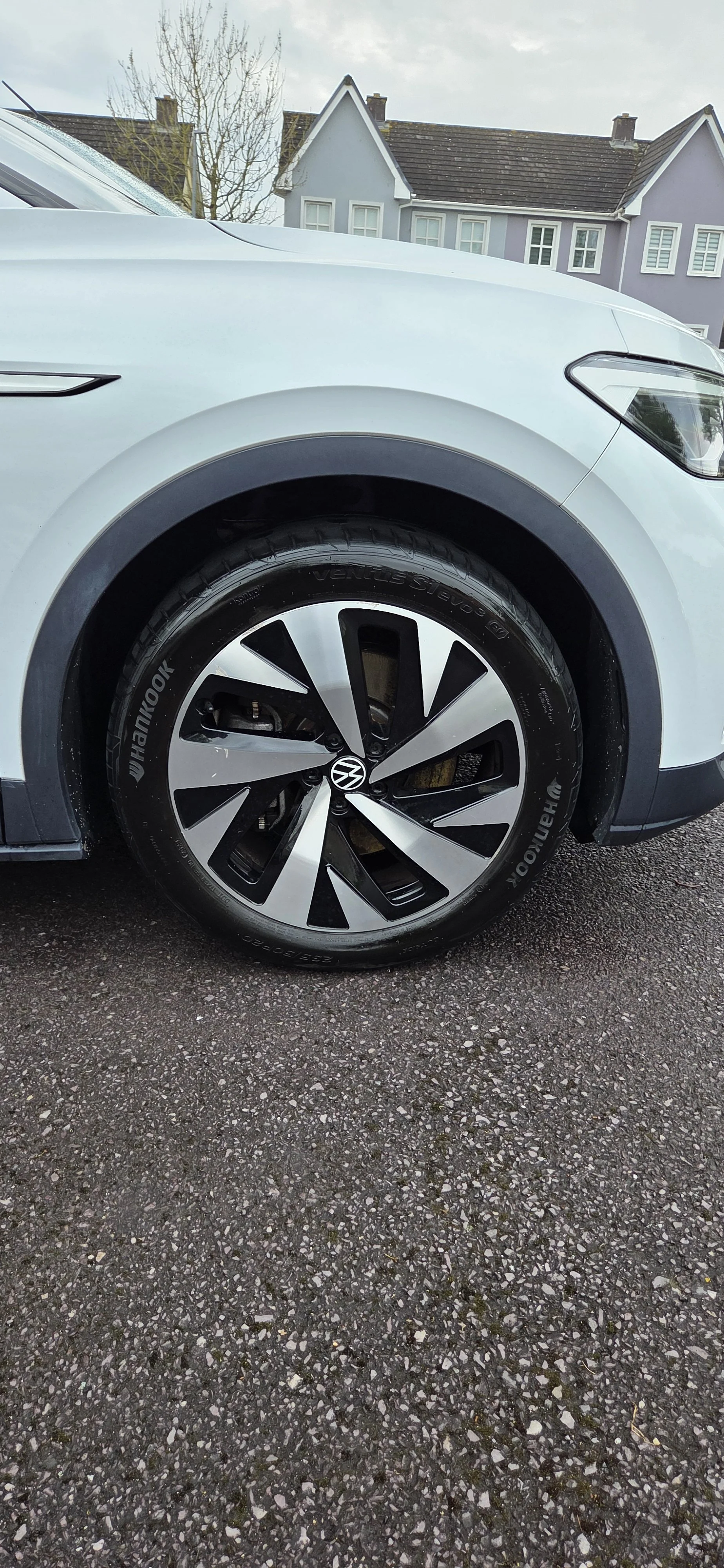 Close-up of the front tire and wheel of a white Volkswagen SUV parked on asphalt with a row of houses in the background.