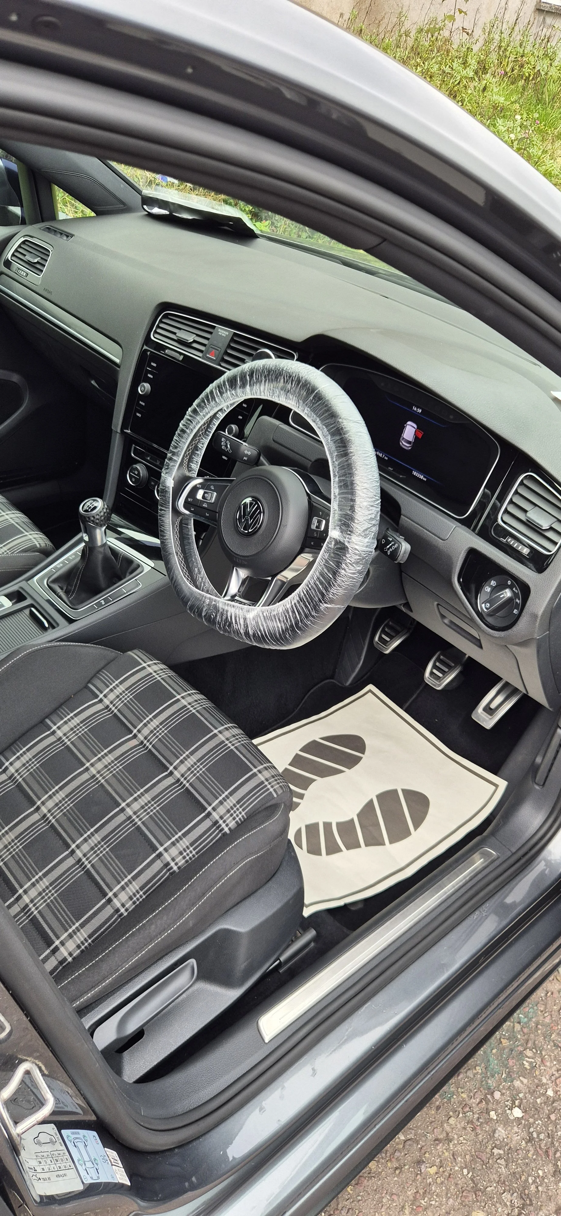 Interior of a Volkswagen car with a plaid-patterned seat, steering wheel with plastic cover, accelerator and brake pedals, and a dashboard display.