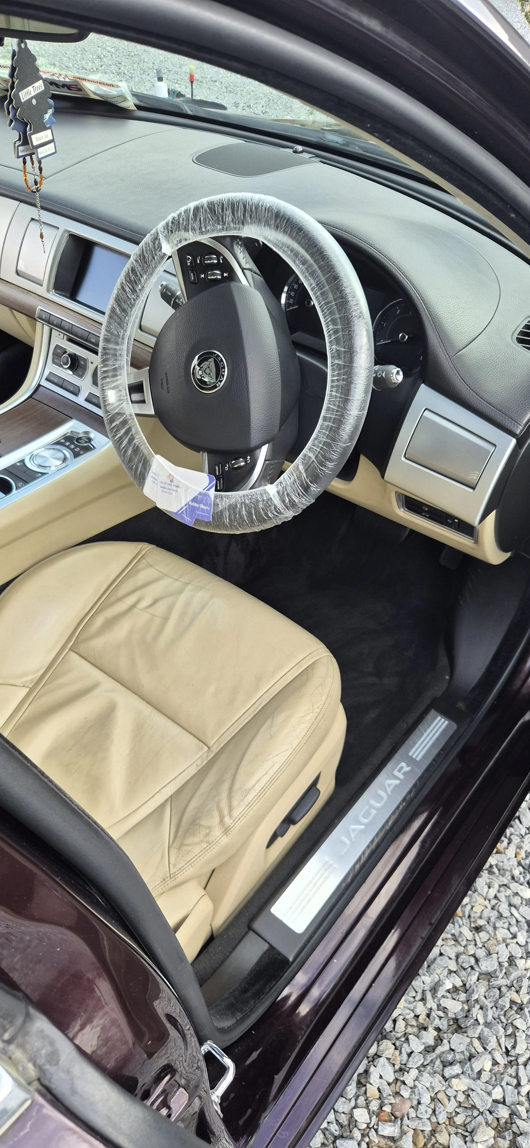 Interior of a Jaguar car, beige leather seat, steering wheel with plastic cover, dashboard with touchscreen, and aftermarket decorations hanging from rearview mirror.