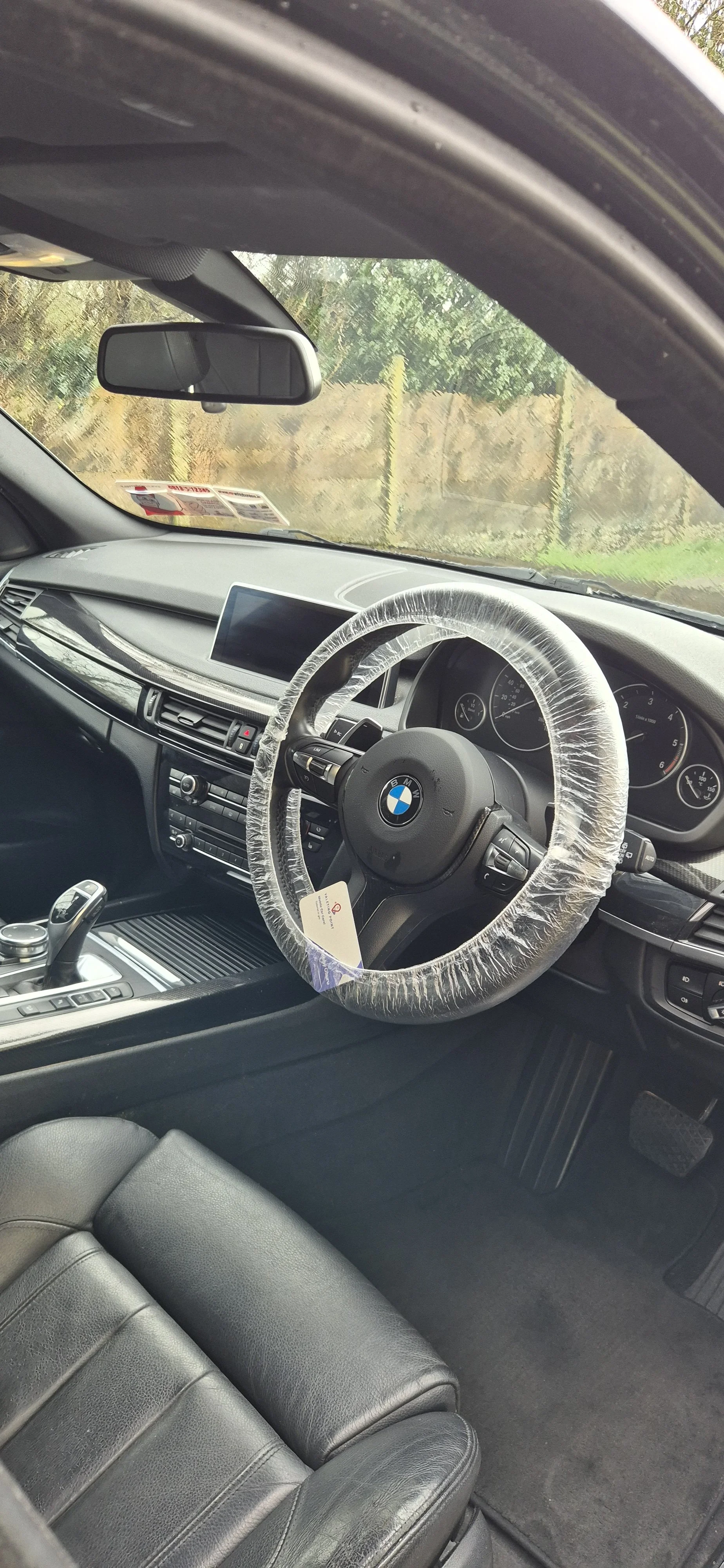 Interior of a BMW car with a plastic-covered steering wheel, black leather seats, dashboard, and gear shifter, parked outdoors.