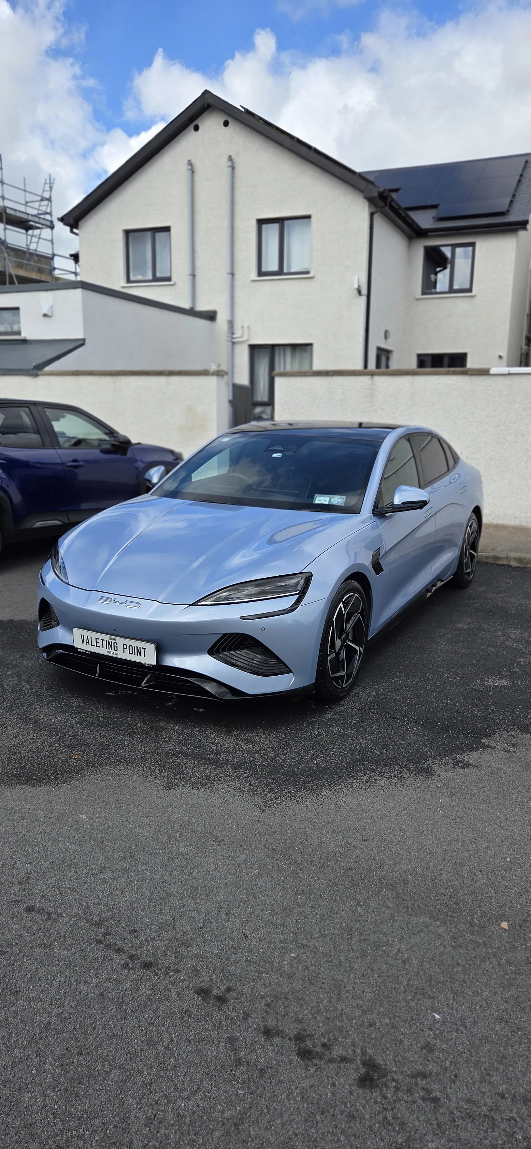 A light blue electric car parked on a paved parking lot in front of residential buildings, with a blue sky and clouds overhead.