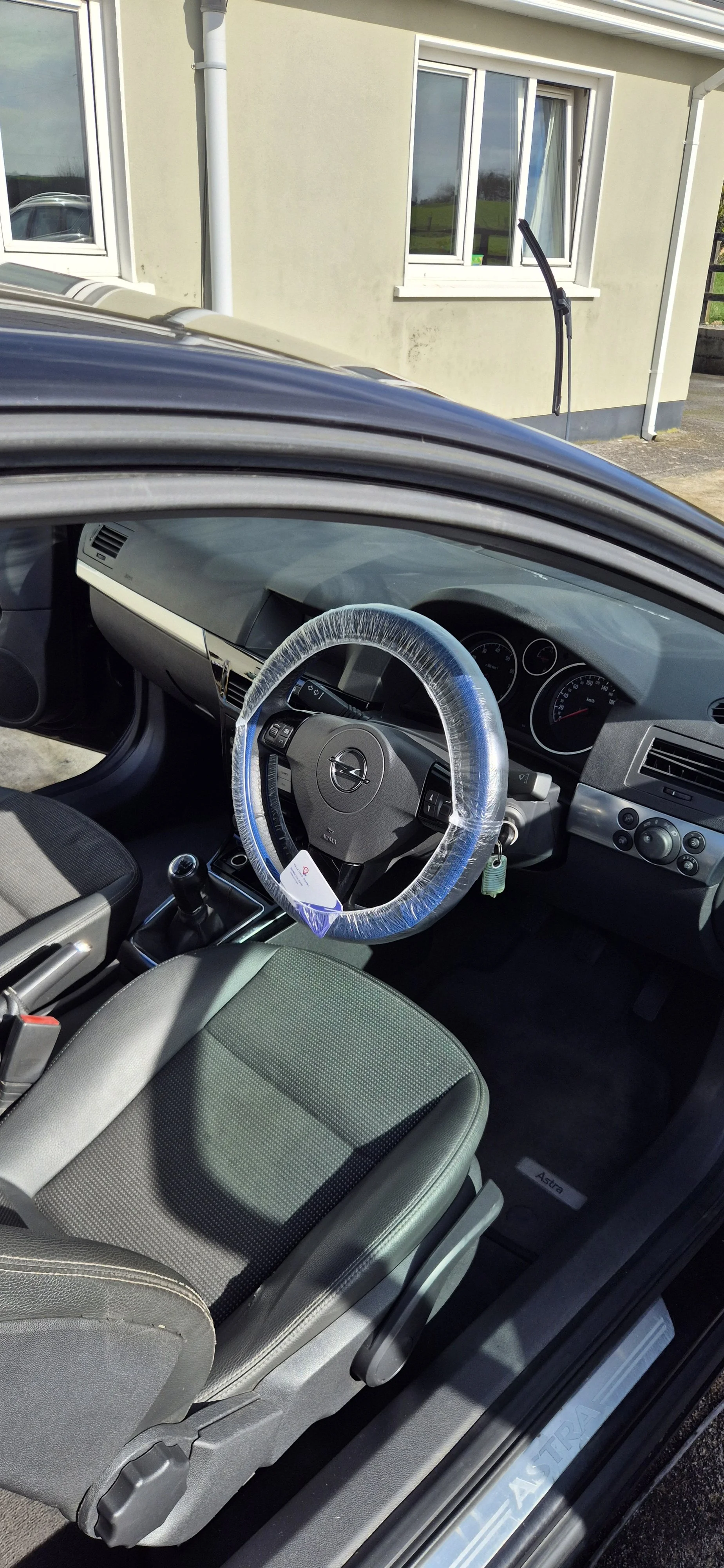 Interior of a car with a plastic-covered steering wheel, manual gear shift, and black fabric seats, parked in front of a house with beige walls and white-framed windows.