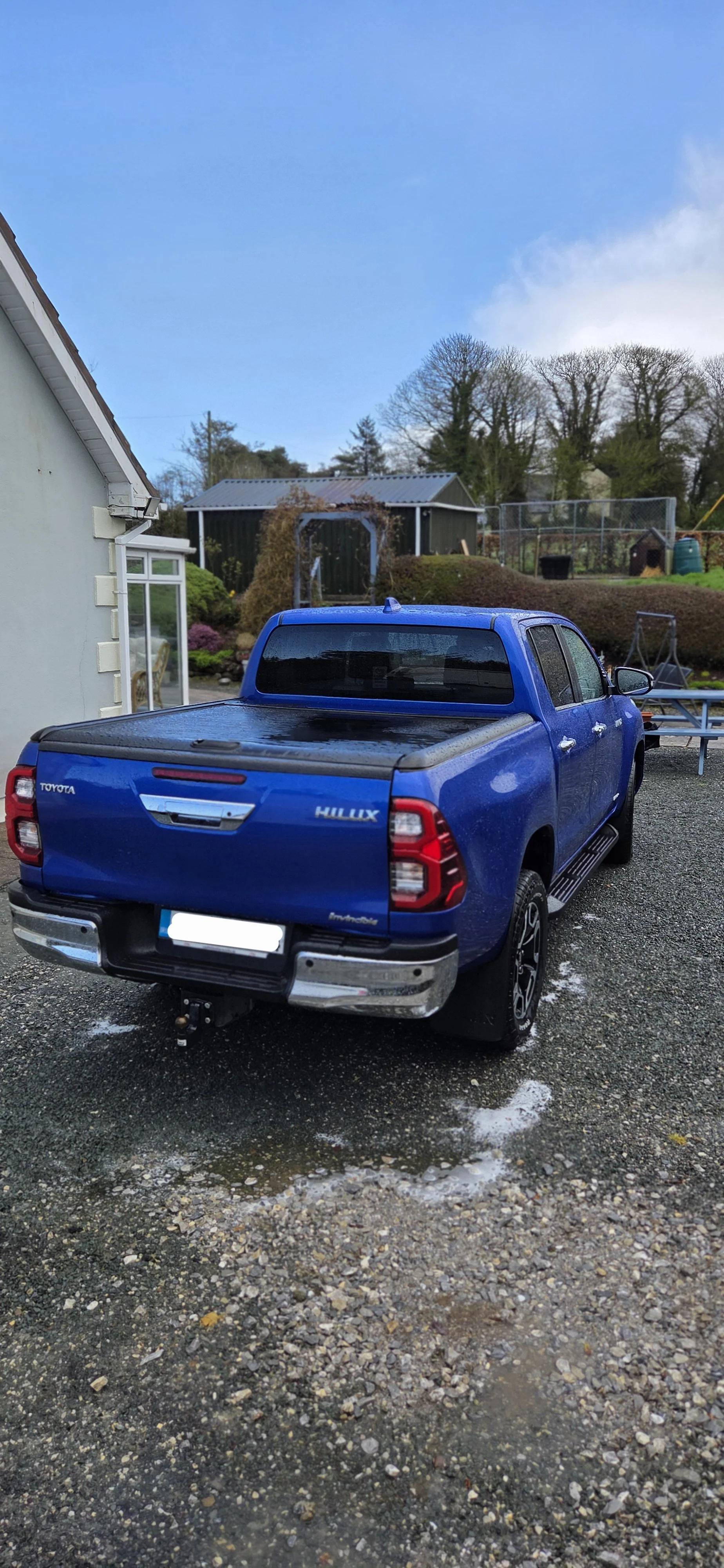 Blue Toyota Hilux pickup truck parked on a gravel driveway with some puddles, beside a light-colored house and a garden with trees and shed in the background. The sky is partly cloudy with patches of blue.