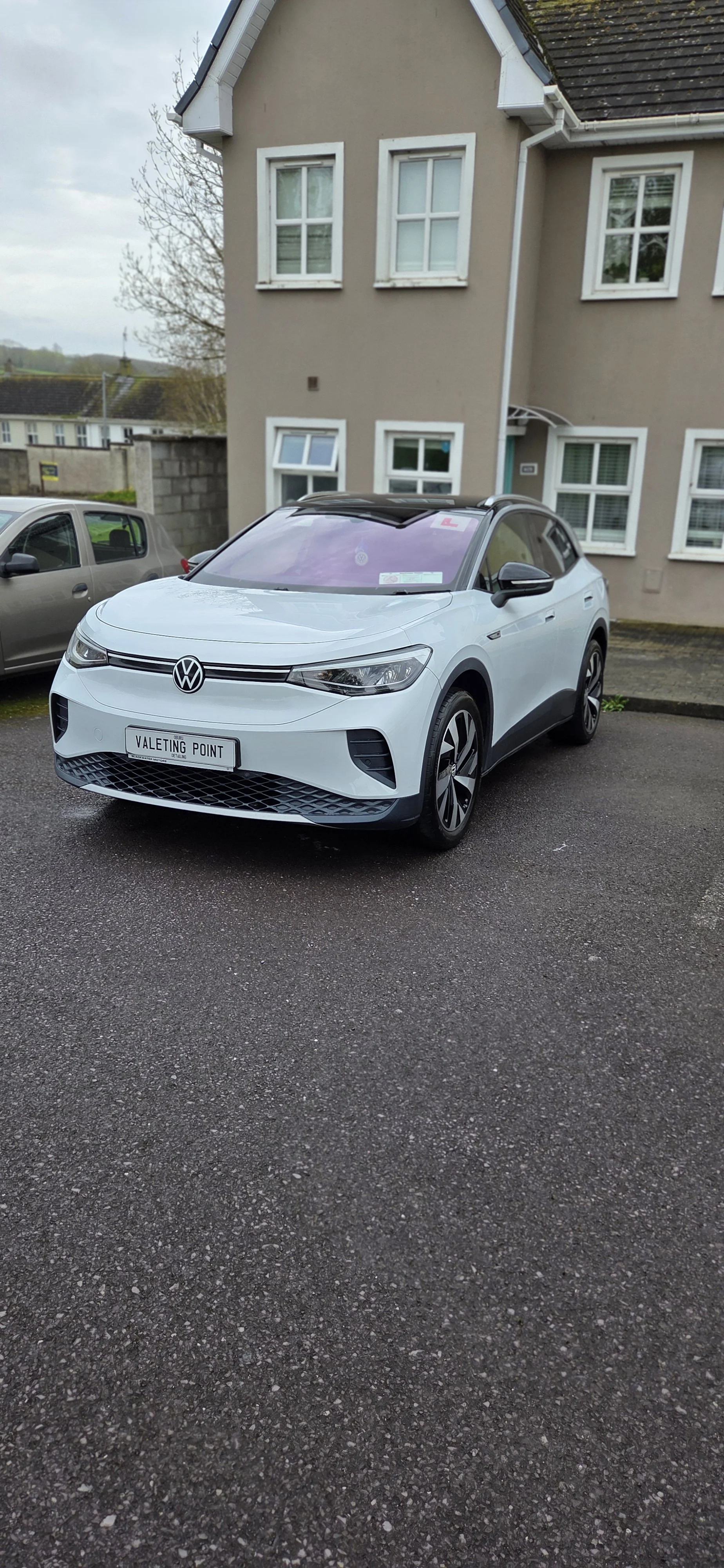 White Volkswagen ID.3 electric car parked in front of a residential building with gray walls and white window frames.