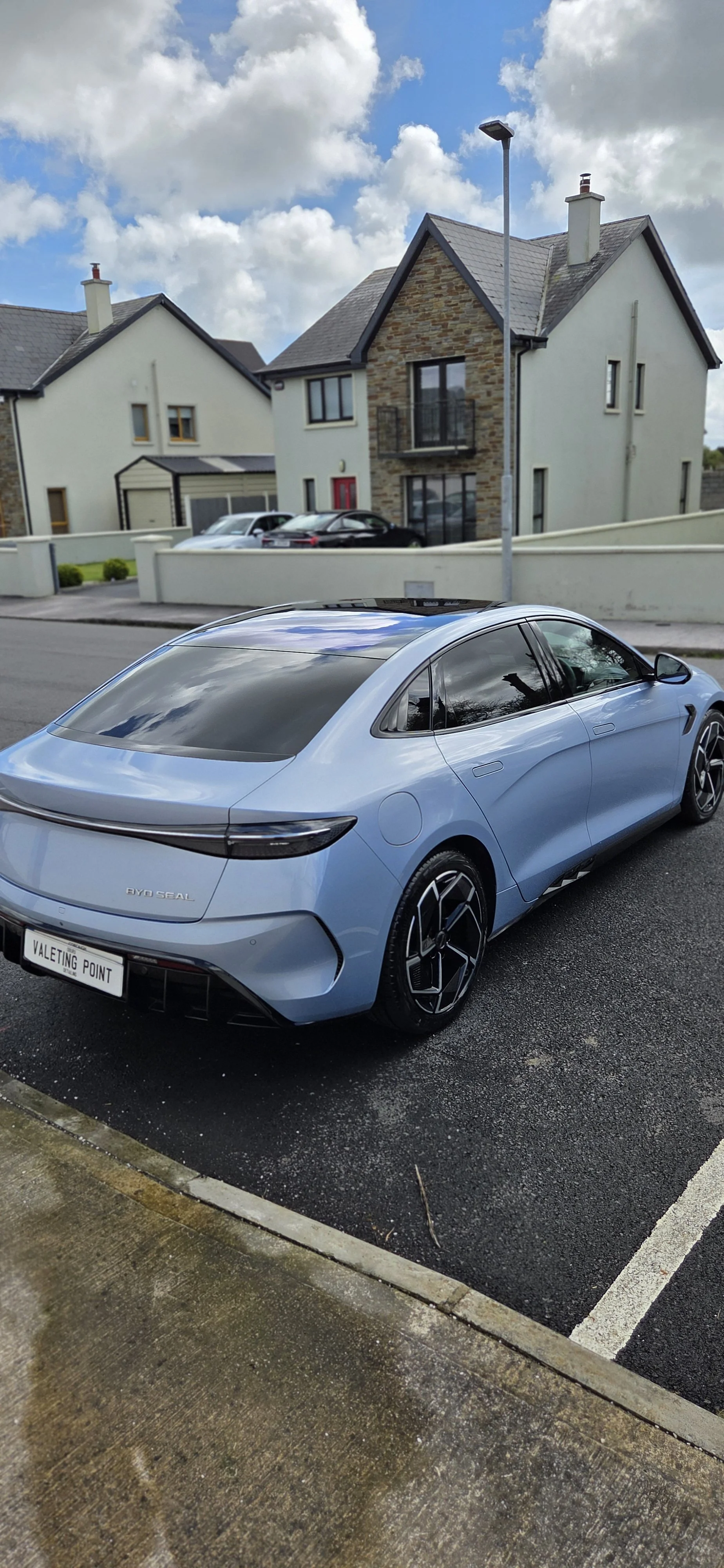 A blue electric car parked on a residential street in front of modern houses under a partly cloudy sky.