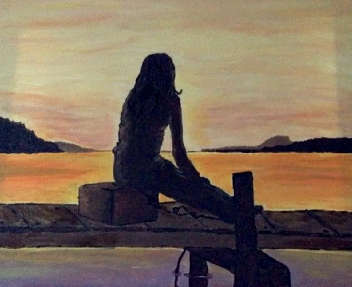 Silhouette of a woman sitting on a dock at sunset, with a body of water and distant landforms in the background.