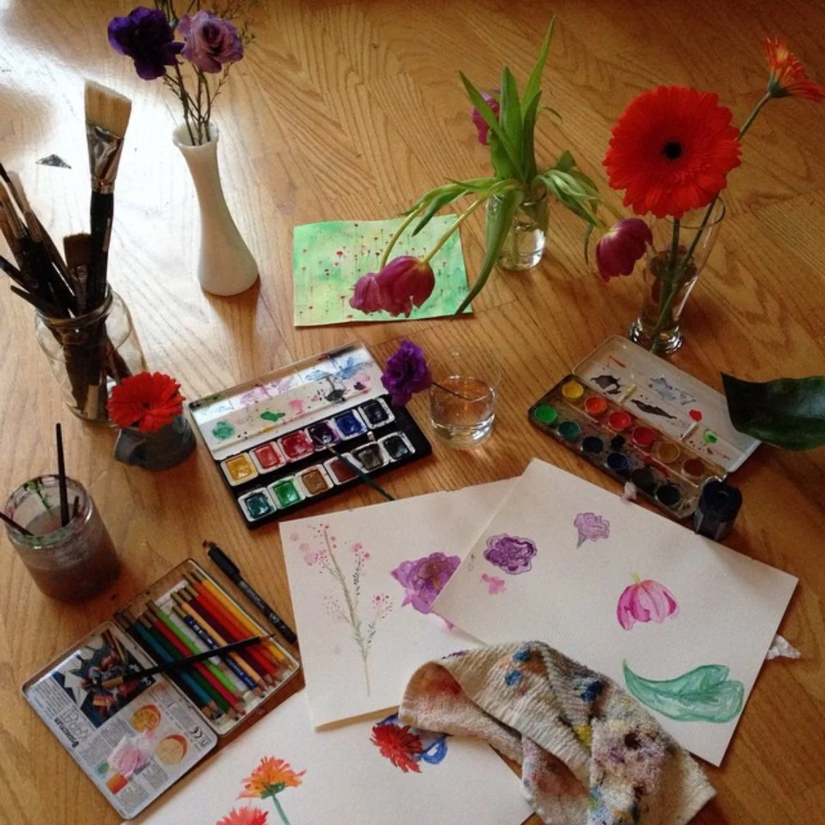 A wooden table with various art supplies, including watercolor paints, colored pencils, and paintbrushes. There are several sheets of paper with watercolor flower paintings, and vases of fresh flowers like poppies, tulips, and other colorful blooms.