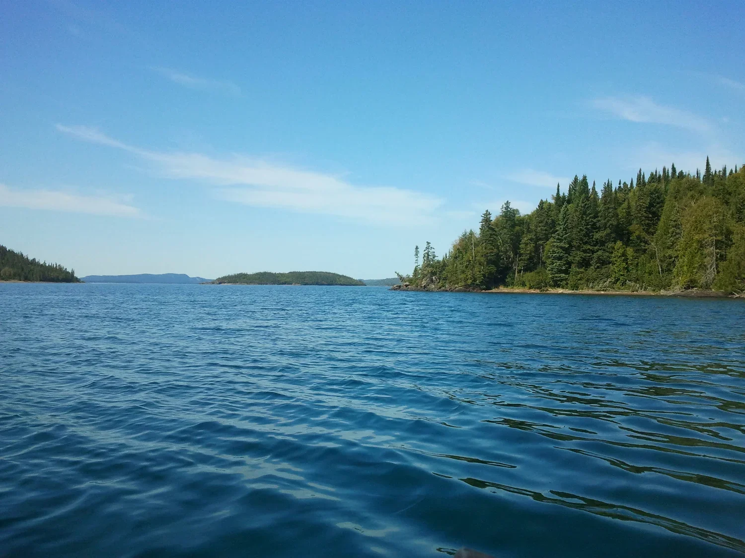 A scenic lake with clear blue water, surrounded by green, forested hills on Nirivia Island under a partly cloudy sky.