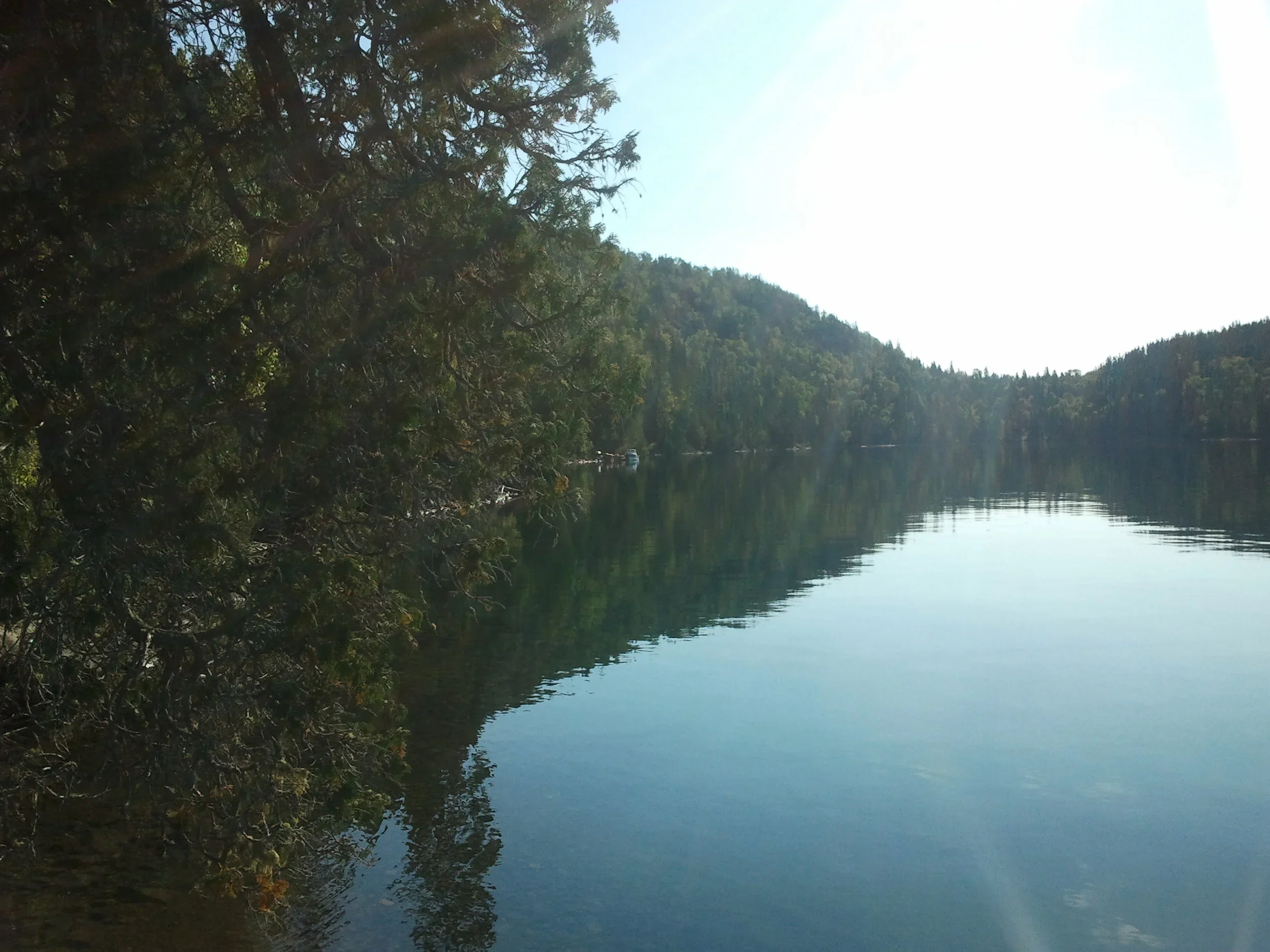 A peaceful lake surrounded by green trees and hills on Nirivia Island, with calm water reflecting the landscape, under a clear sky.