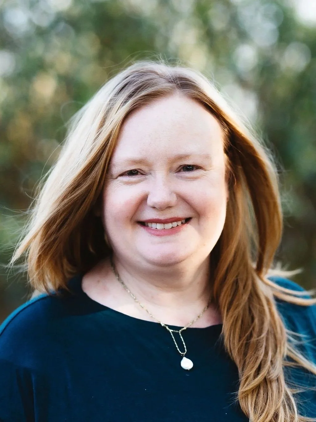 Close-up of art therapist Cassandra Evans with long red hair smiling outdoors, wearing a black top and a pearl necklace.