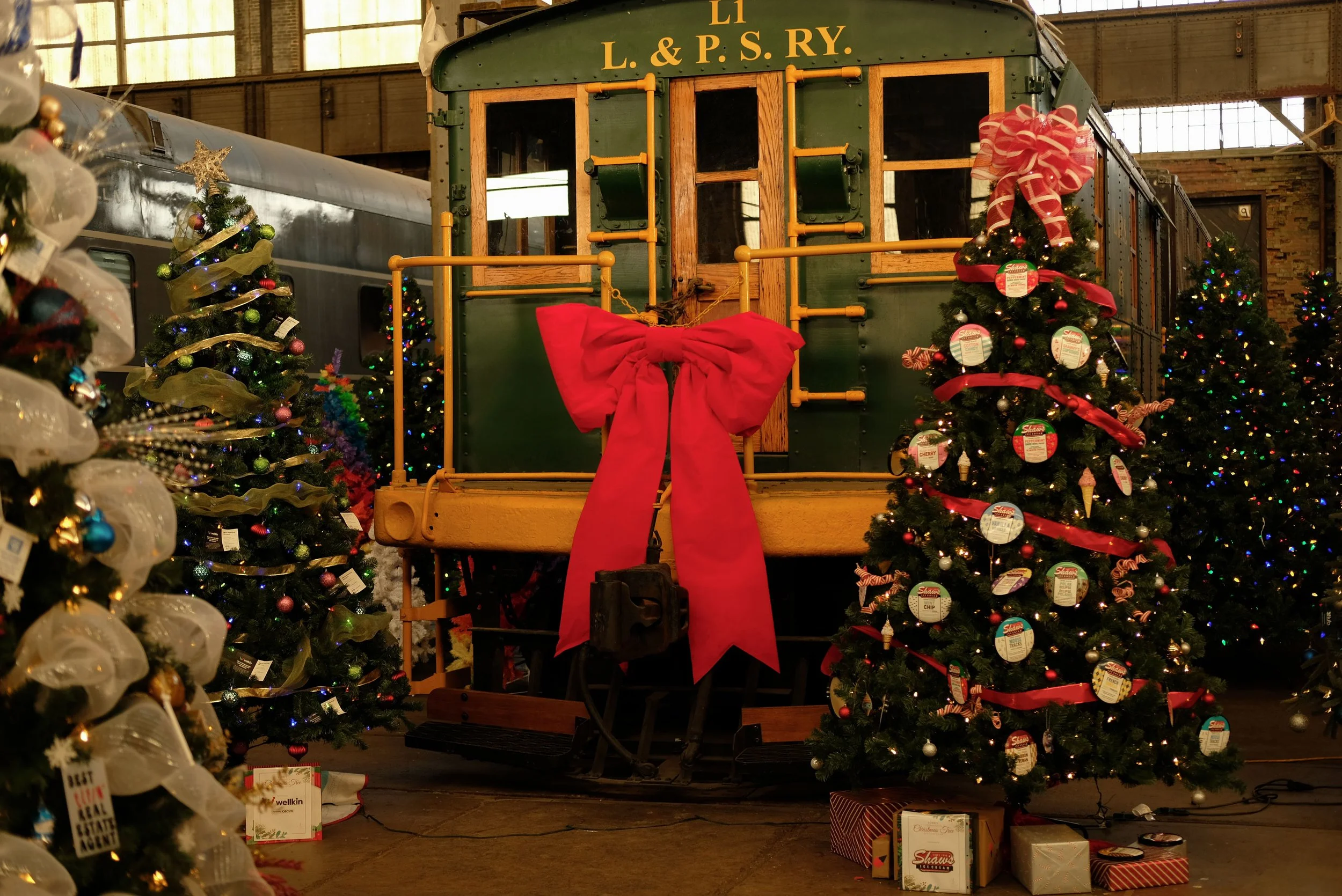 Display of vintage train car decorated for Christmas with a large red bow. Christmas trees adorned with ornaments and lights are on both sides of the train car.