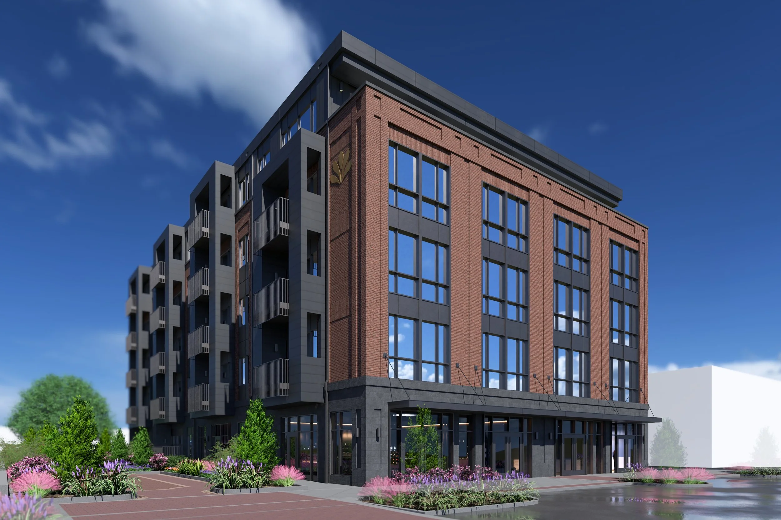 A modern multi-story building with large windows, brick and dark panel exterior, surrounded by landscaped greenery and pink flowering bushes against a clear blue sky.