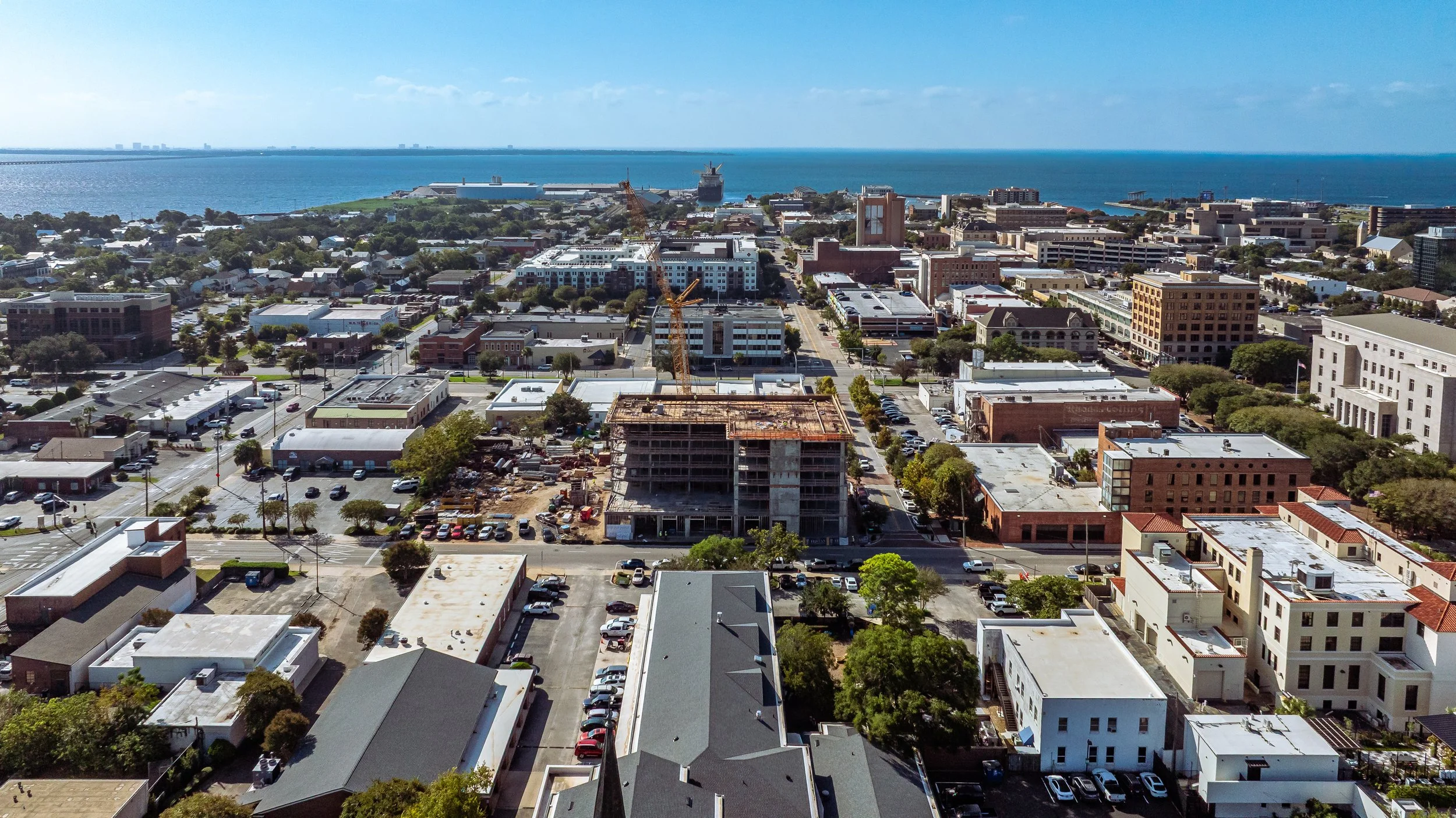 Aerial view of a city near the coast with buildings, streets, and a construction site with cranes, overlooking the ocean