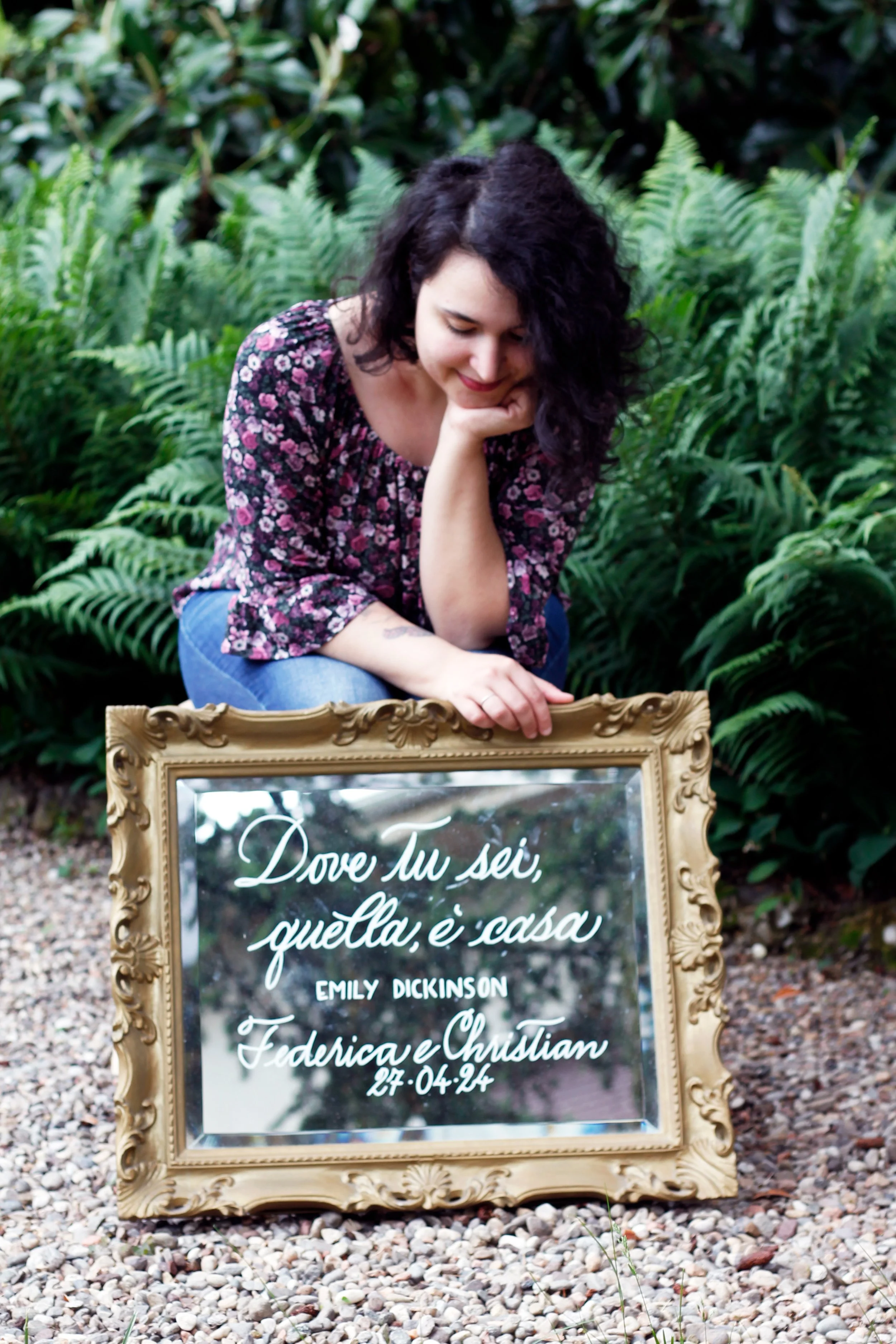 Una donna con capelli ricci neri indossa una camicia a maniche lunghe con stampa floreale e jeans, si china su un'installazione di un quadro decorativo all'aperto, circondata da piante verdi e foglie di felce.