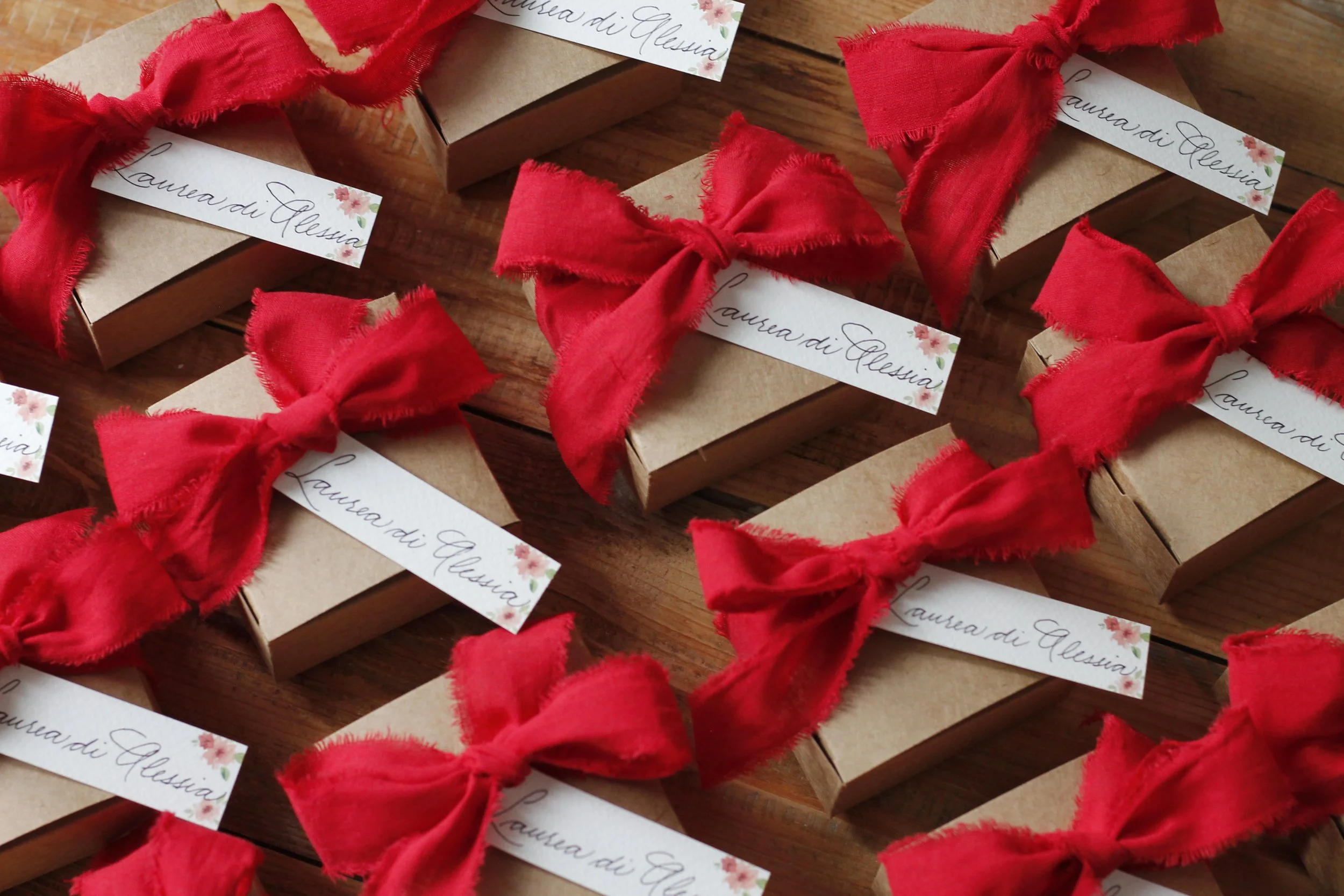 Piccoli regali confezionati in scatola di cartone marrone, decorati con nastri di stoffa rossa e etichette con scritto 'Laurea di Alessia' con piccole decorazioni floreali.