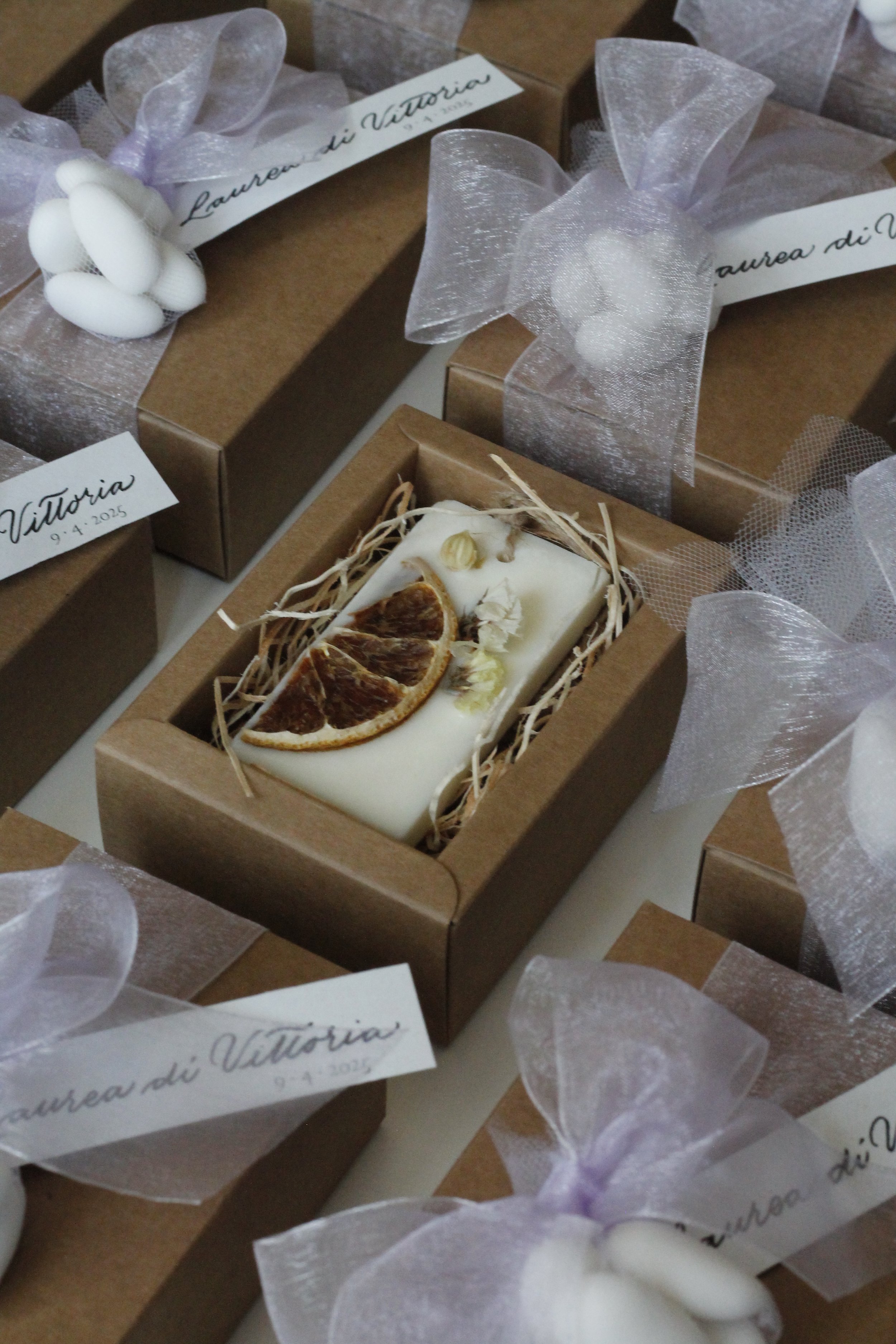 Sapone artigianale decorato con scorze di limone e fiori secchi, confezionato in una scatola di cartone, circondato da confezioni regalo con nastro di organza e etichette con scritto ' Laurea di Vittoria'.
