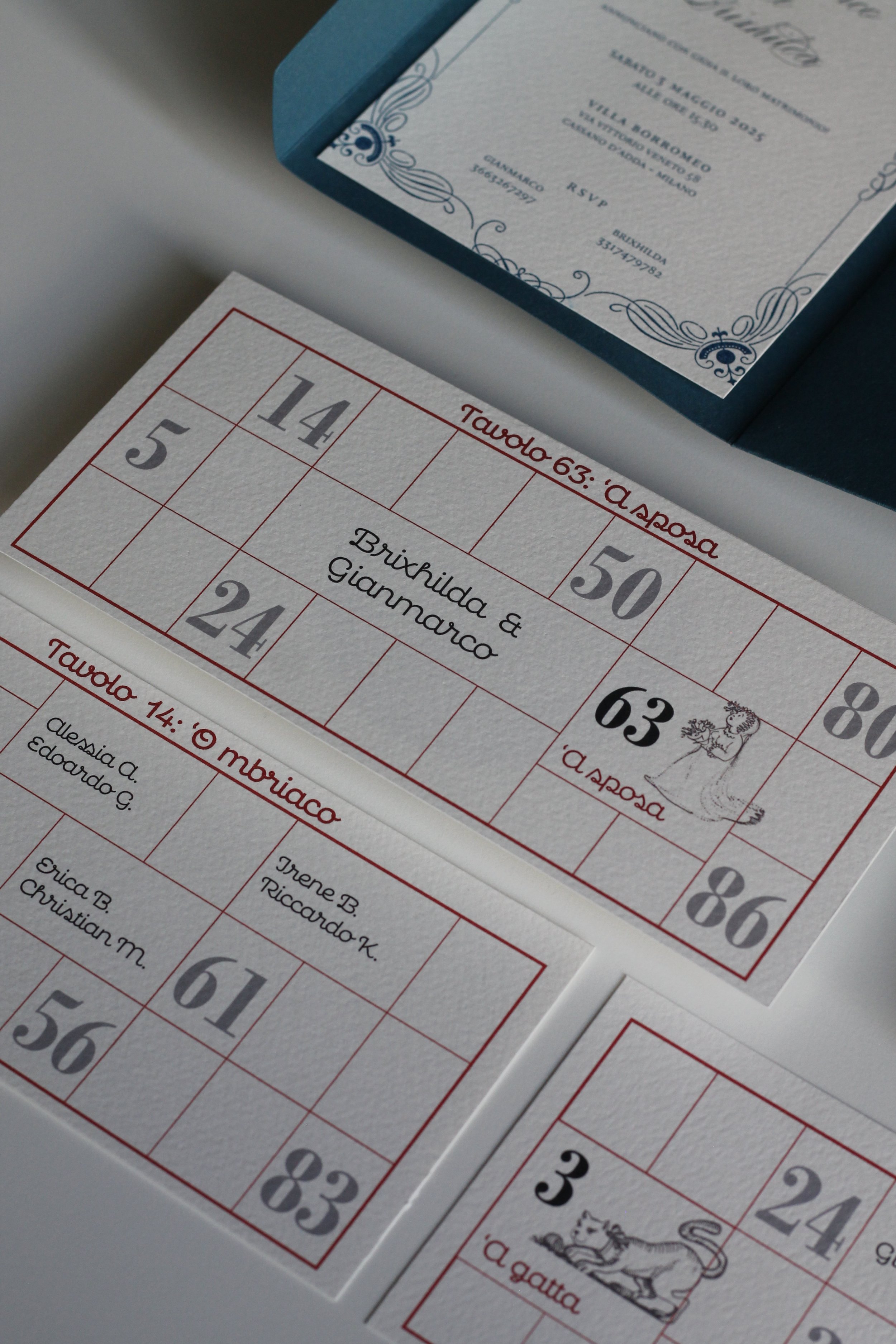 Cartella di giochi numerici con numeri e nomi di coppie di sposi, accompagnata da un'invito formalizzato in un elegante biglietto bianco e blu.