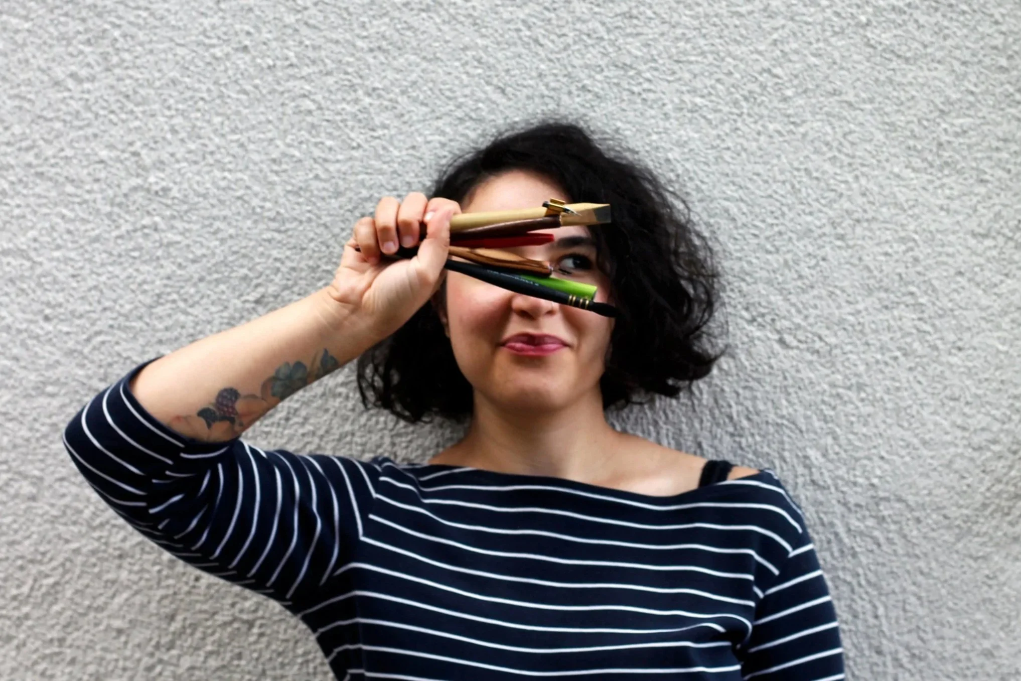 Una donna con capelli ricci e tatuaggi sull'avambraccio tiene in mano vari colori di penne e le brandisce davanti a un muro di pietra beige.