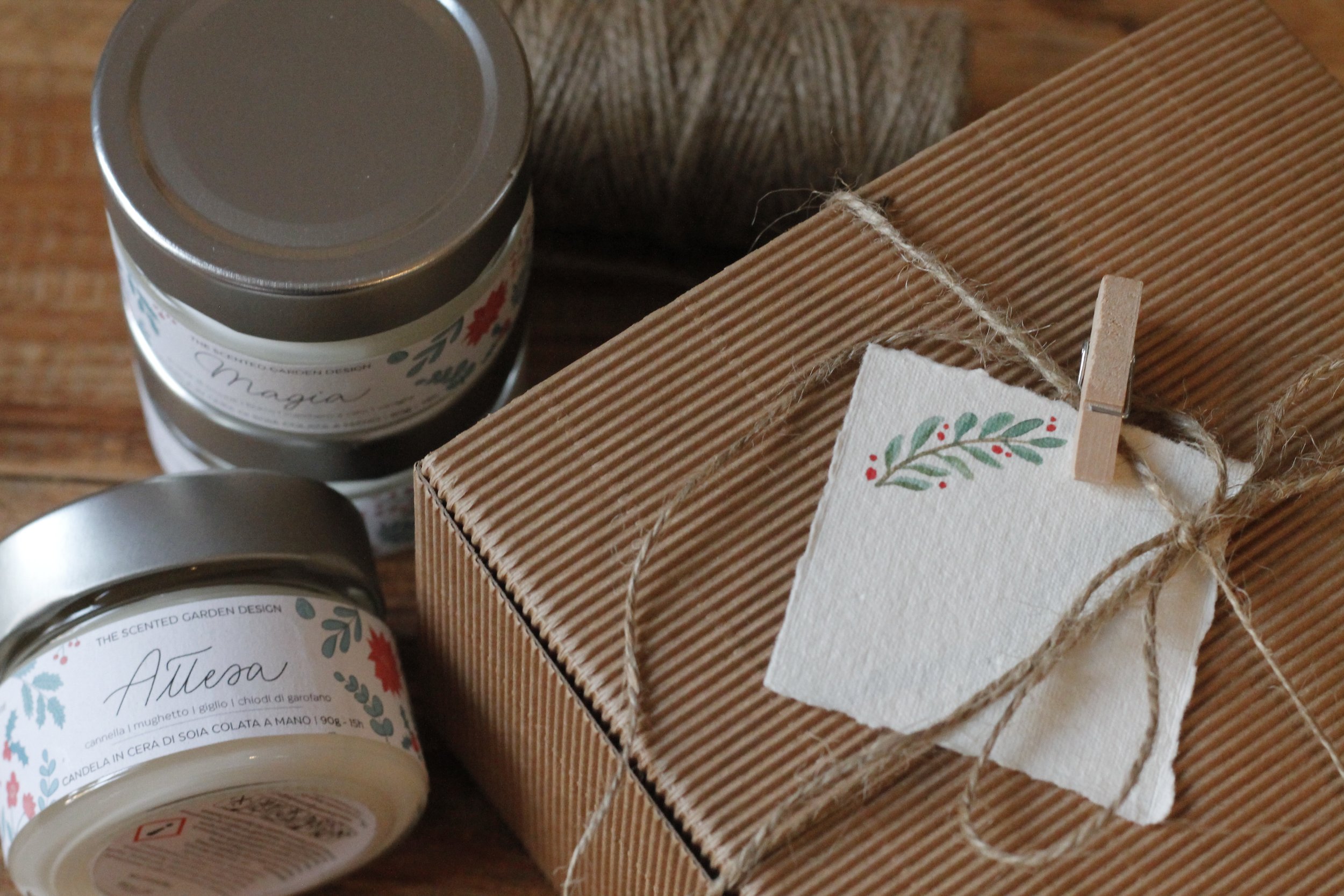 Pacchetto regalo di carta canasta, due barattoli di cera profumata con etichette decorate con motivi floreali, su superfice di legno.