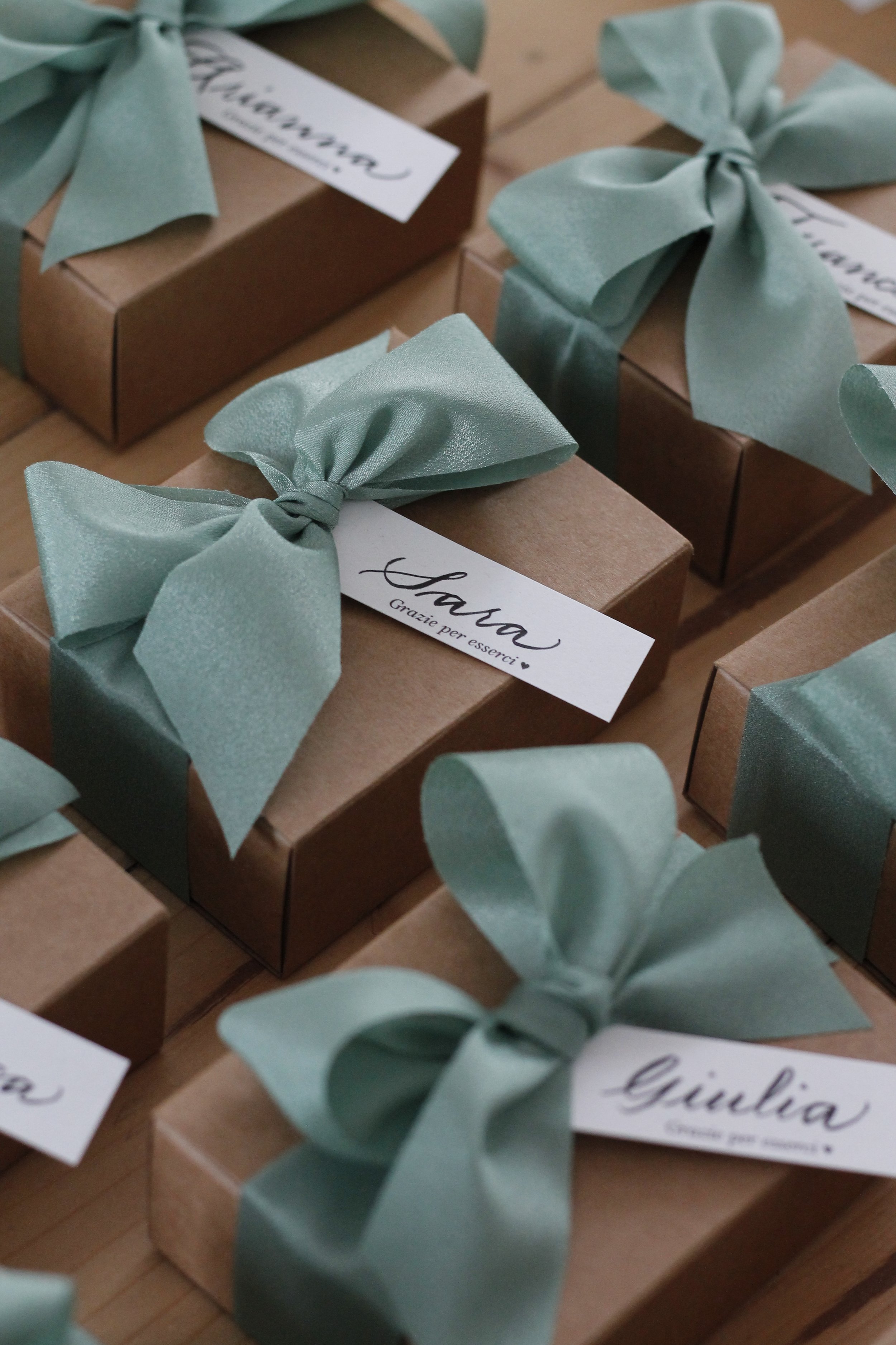 Confezioni regalo di cartone marrone decorate con fiocchi di raso verde acqua e con etichette bianche con scritte in caratteri eleganti.