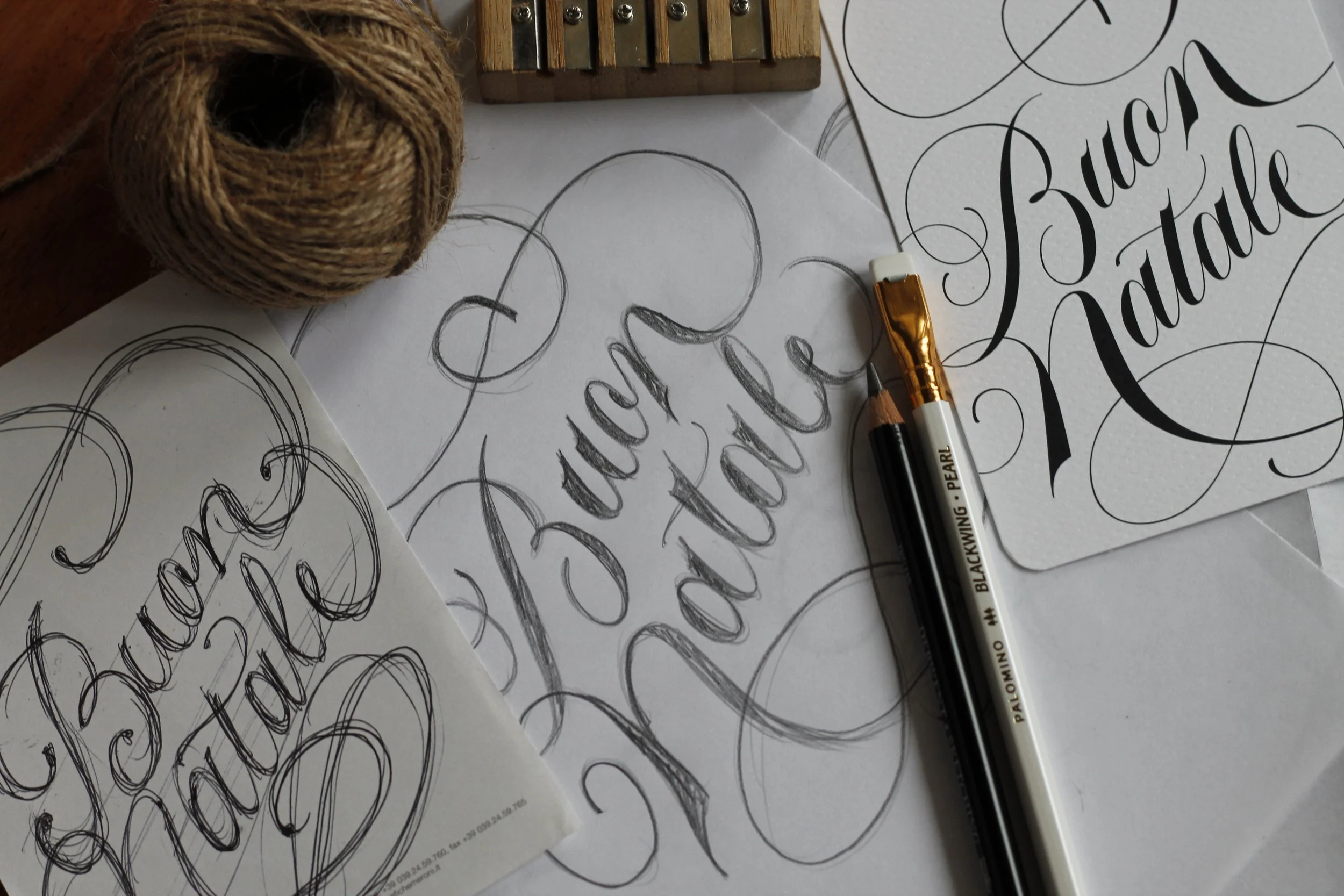 Progetti di calligrafia con parole come 'Buon Natale' e 'Buona Notte', matite, penne e fogli con schizzi e scritte in stile calligrafico, fogli con scritte elaborate, bobina di spago, tavolo di lavoro.