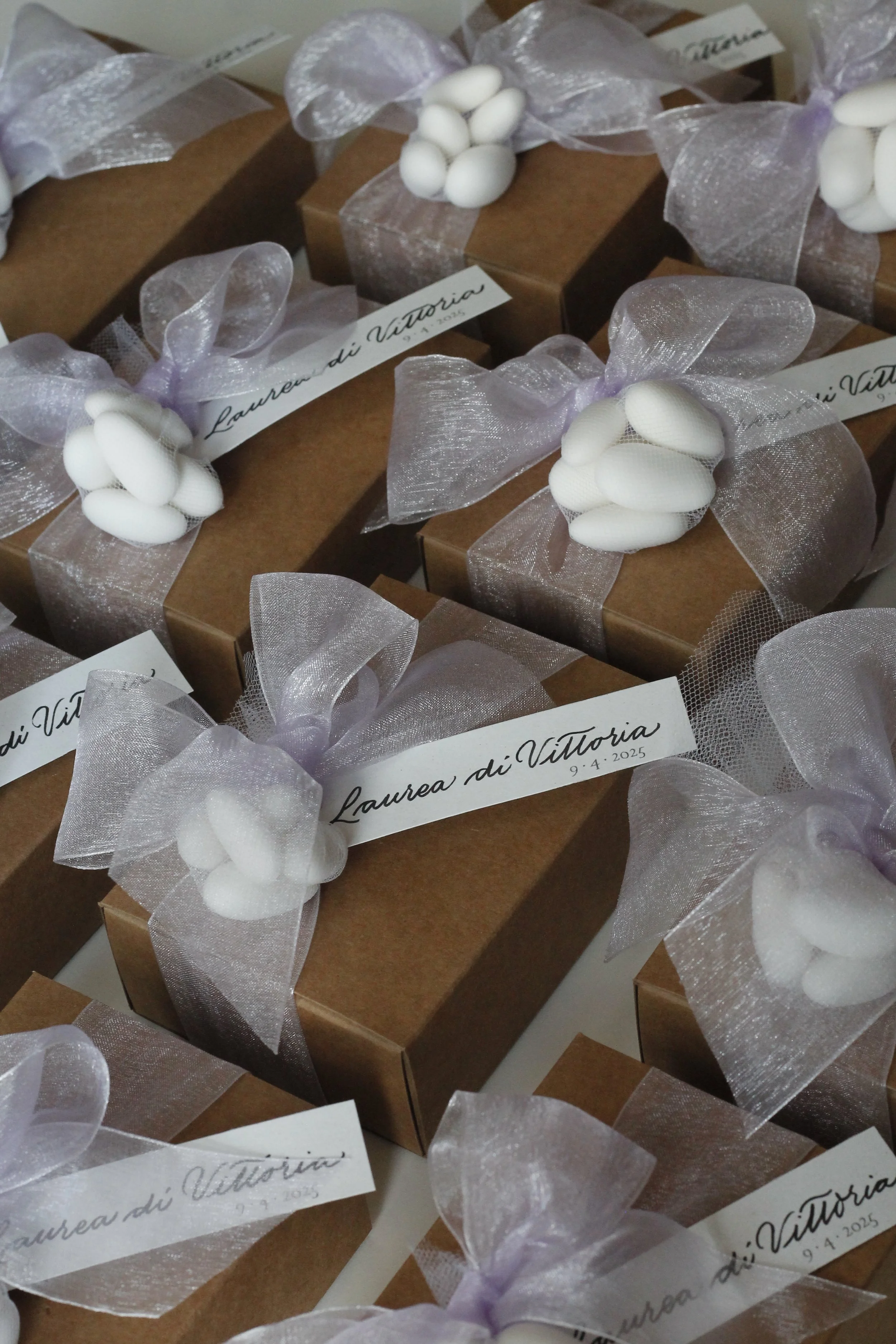 Confezioni regalo di colore marrone con fiocchi di tessuto trasparente, decorati con piccoli rami di ovetti bianchi e biglietti con la scritta 'Laurea di Vittoria' e la data 9.4.2025.