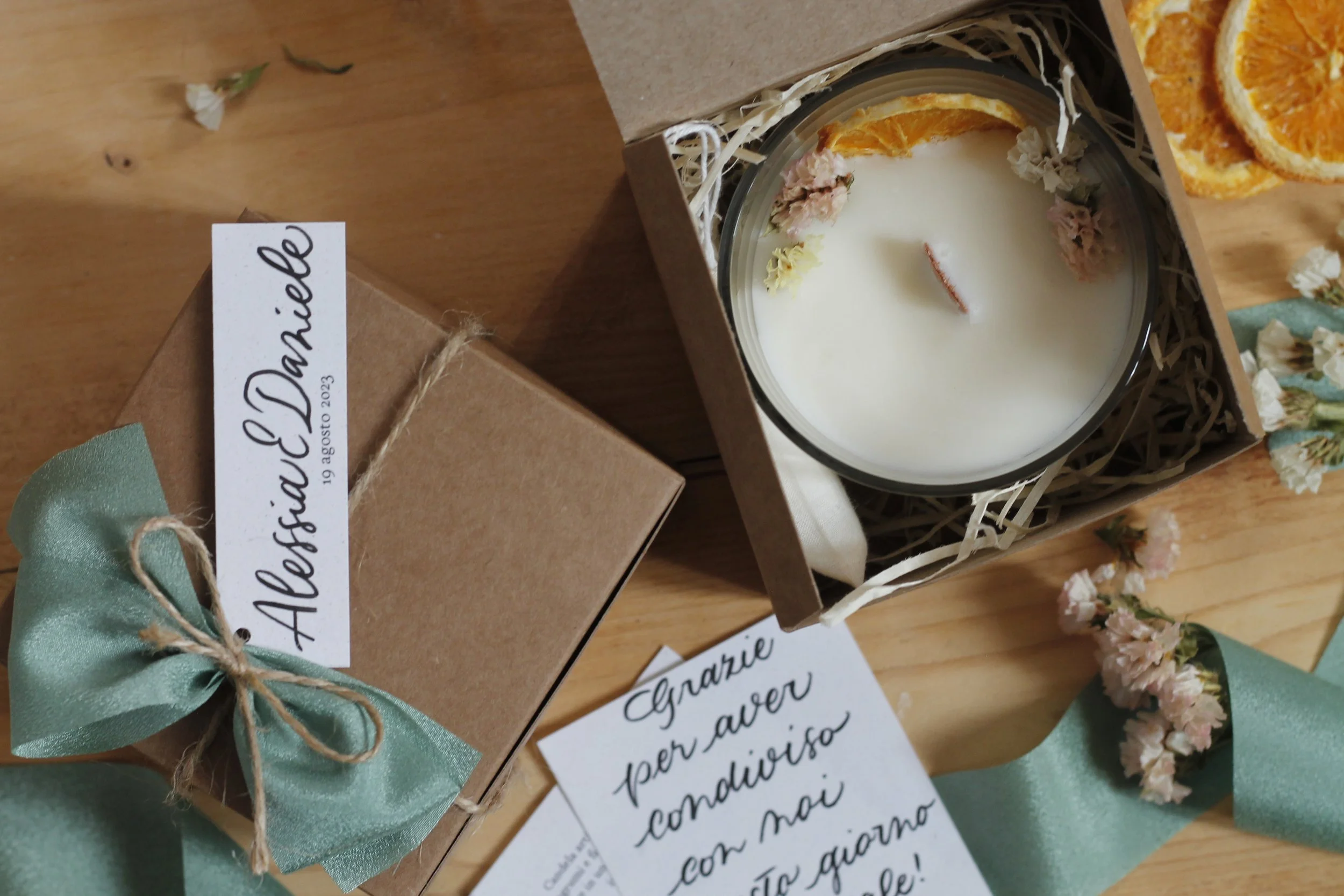Candela in una scatola di cartone decorata con un nastro di raso verde, accompagnata da fiori e note di ringraziamento scritte a mano.