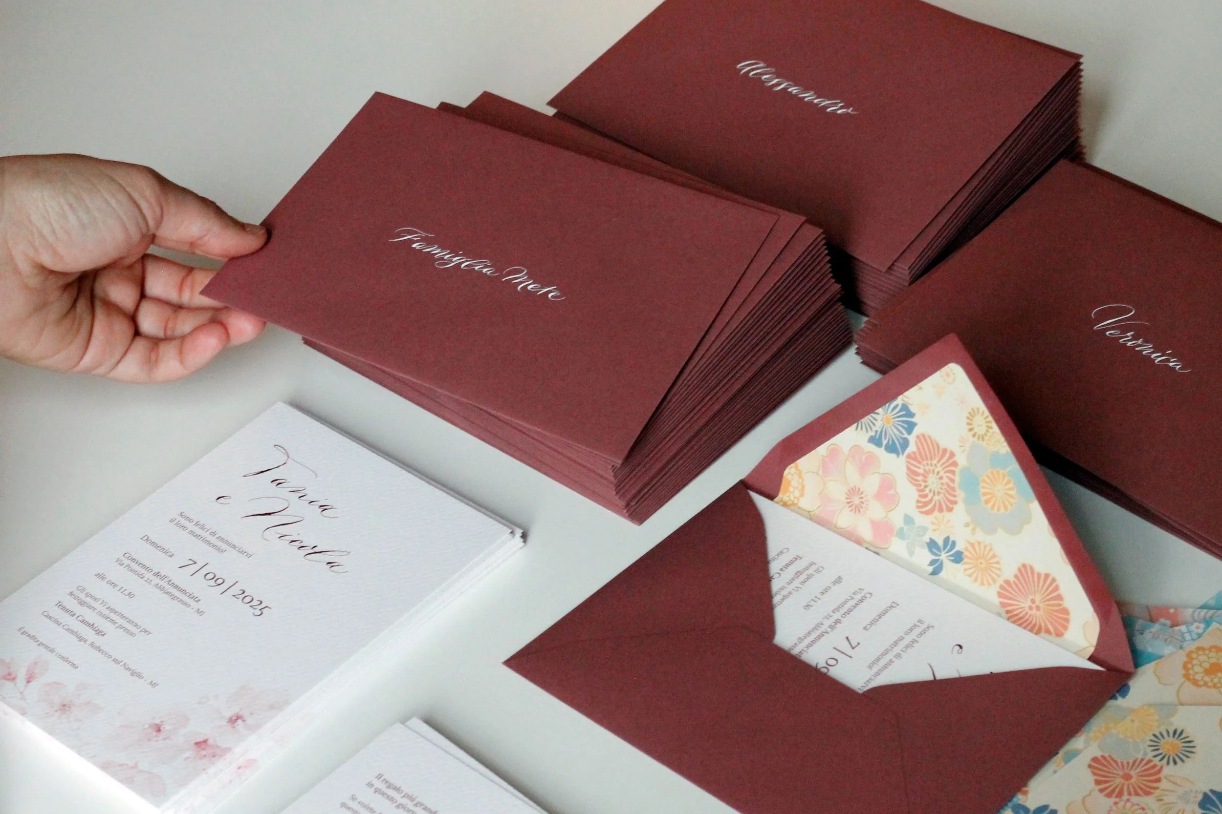 Biglietti di invito color ciliegia con scritte calligrafiche e volantini con decorazioni floreali colorate.