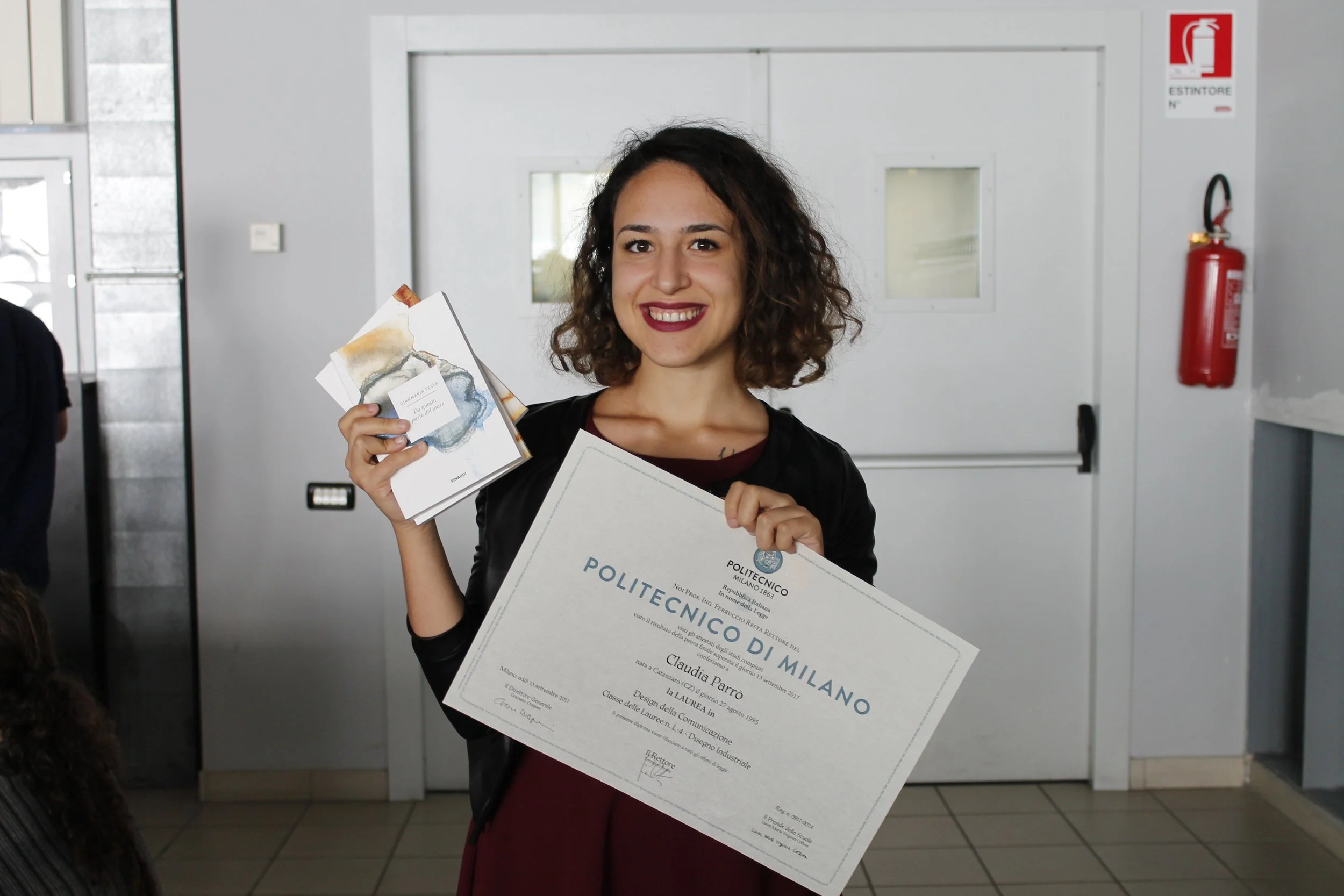 Una donna sorridente tiene in mano un diploma di laurea dell'Università Politecnico di Milano e alcuni opuscoli, in un'area interna di un edificio.