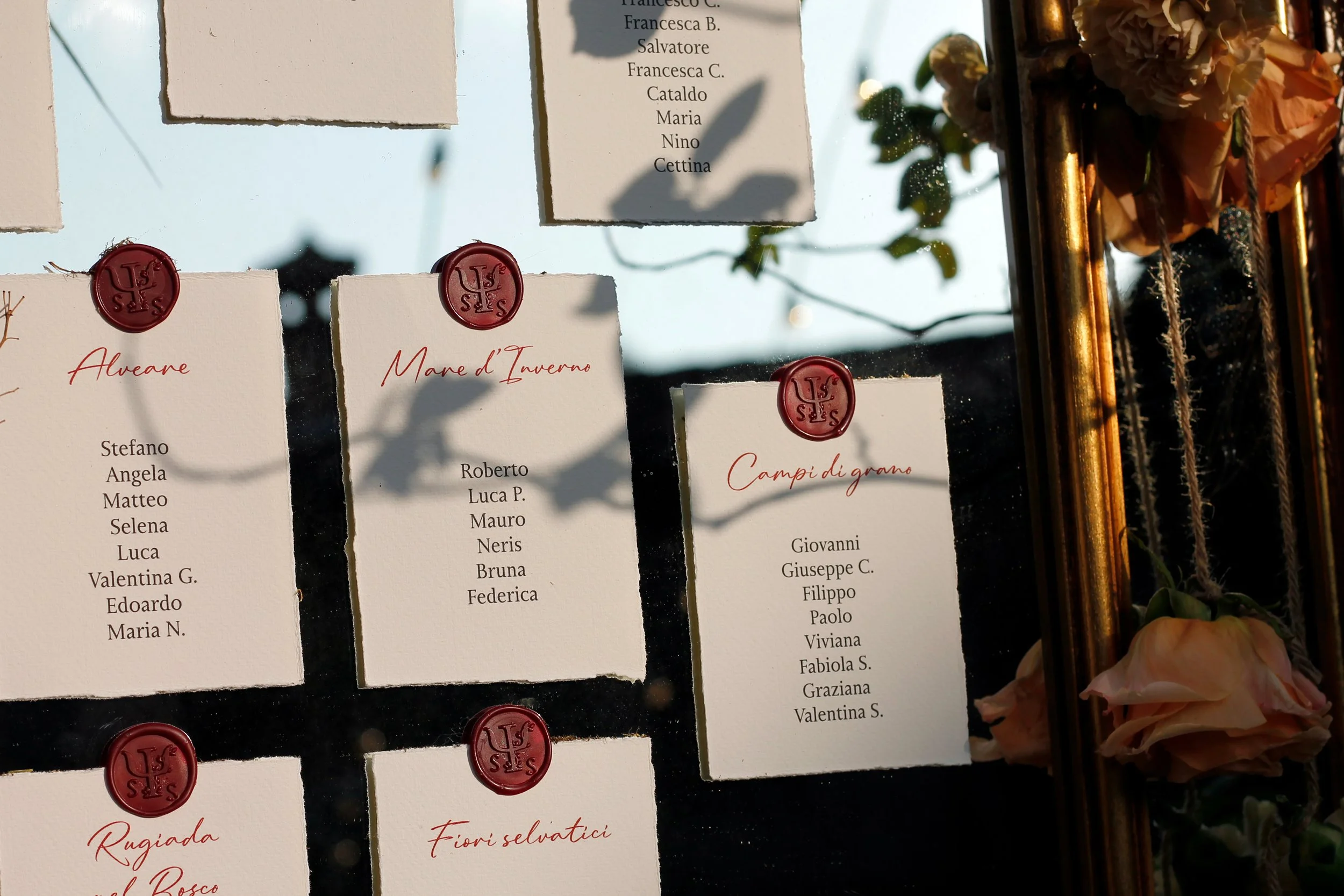 Tabelloni con nomi scritti a mano e sigillo di cera rossa, con decorazioni floreali e foglie verdi.
