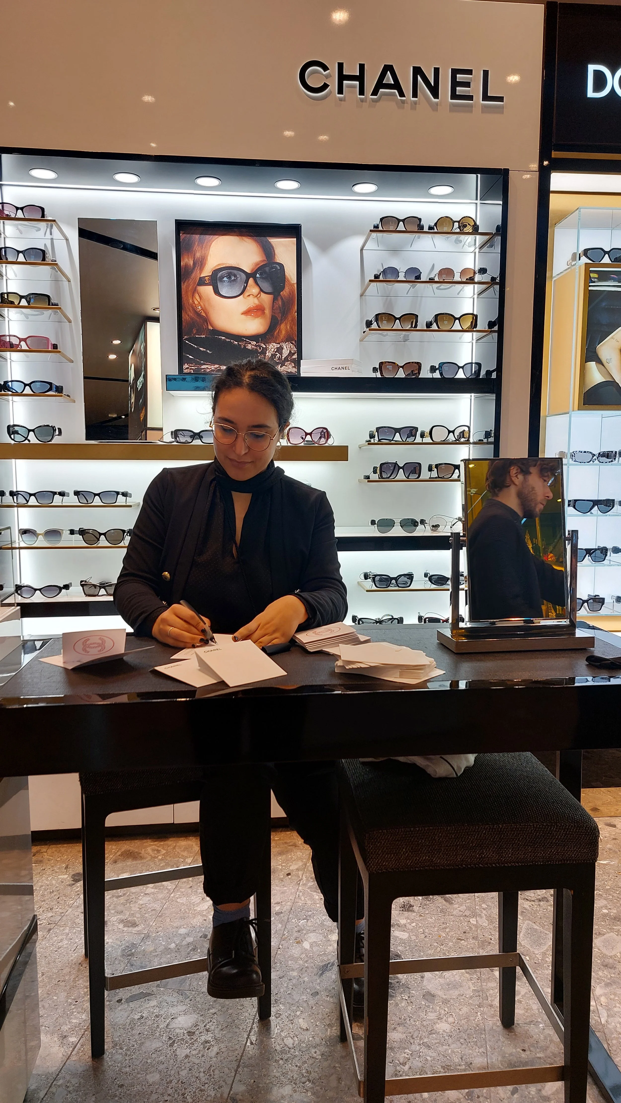 Interno di un negozio di occhiali di marca Chanel, con scaffali pieni di occhiali da sole e da vista. Una donna con occhiali è seduta dietro un bancone che sta facendo dei fogli, davanti a un grande specchio.