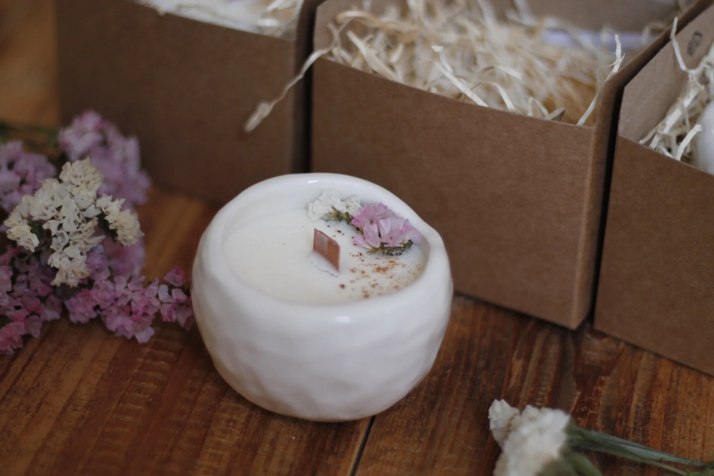 Una candela bianca decorata con fiori viola e una rosa sulla parte superiore, circondata da fiori secchi e scatole di cartone con paglia.